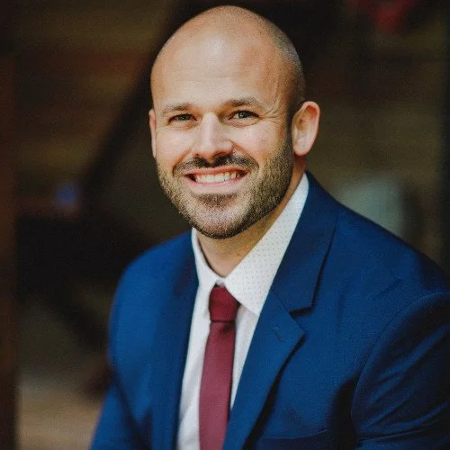 A smiling man with a beard and bald head, wearing a navy suit, white shirt, and maroon tie, in a professional setting.