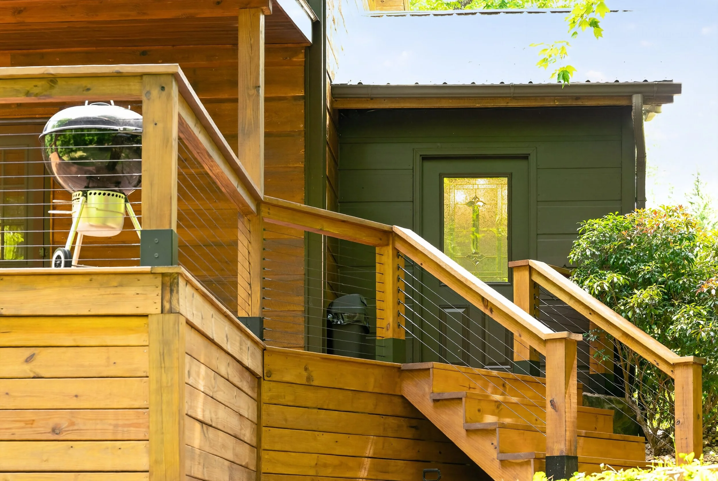 View of a wooden deck with stairs, a fenced railing, a black and chrome grill, and a black door with a glass window reflecting sunlight. Green foliage in the background and a partly cloudy sky.