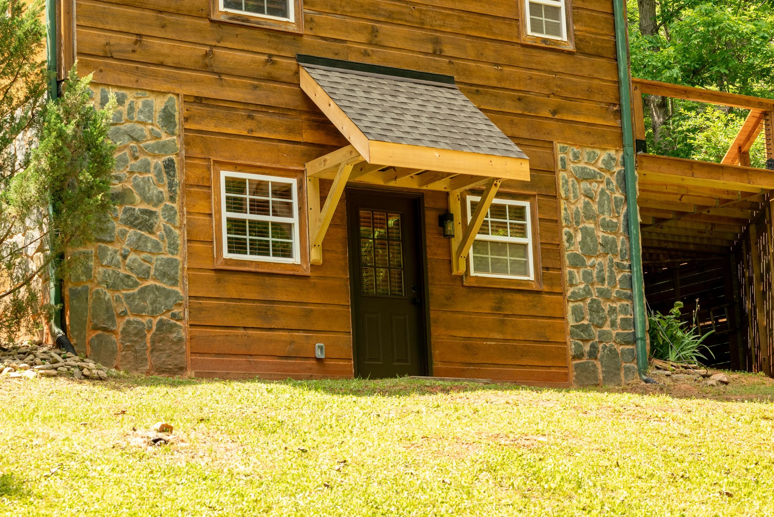 A rustic house with wooden siding and stone accents, with a small covered porch, surrounded by greenery and trees.