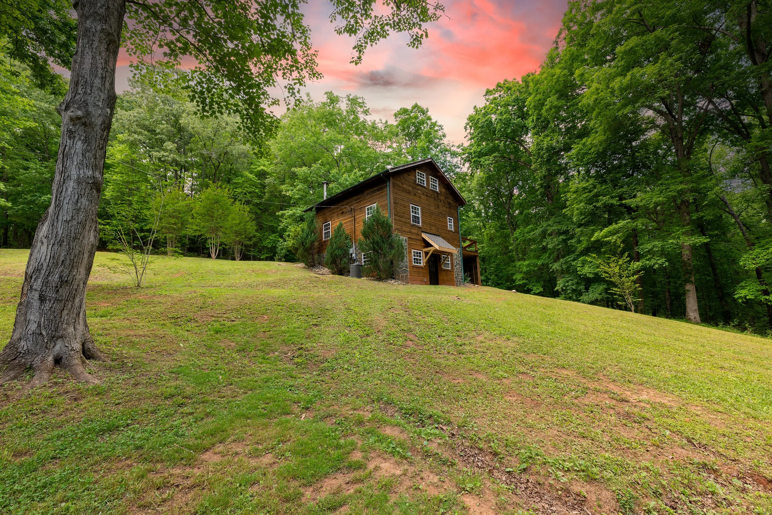 A wooden house with a stone chimney, surrounded by green trees and grass, under a pink and orange sky at sunset.