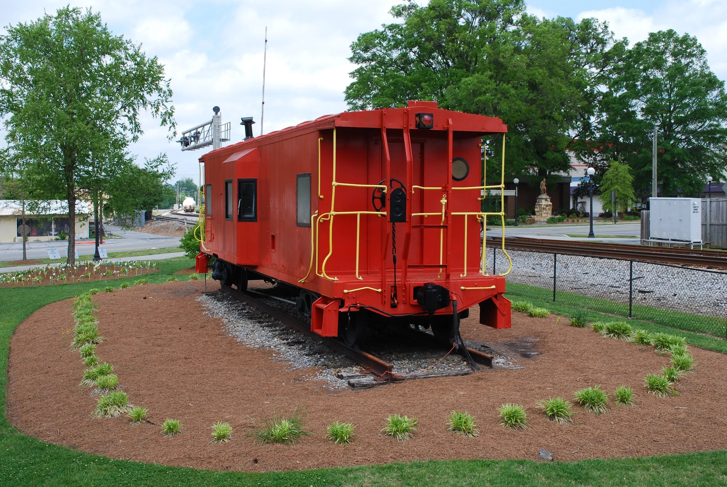 A vintage red train car on display outdoors with green trees, a building, and a street in the background.