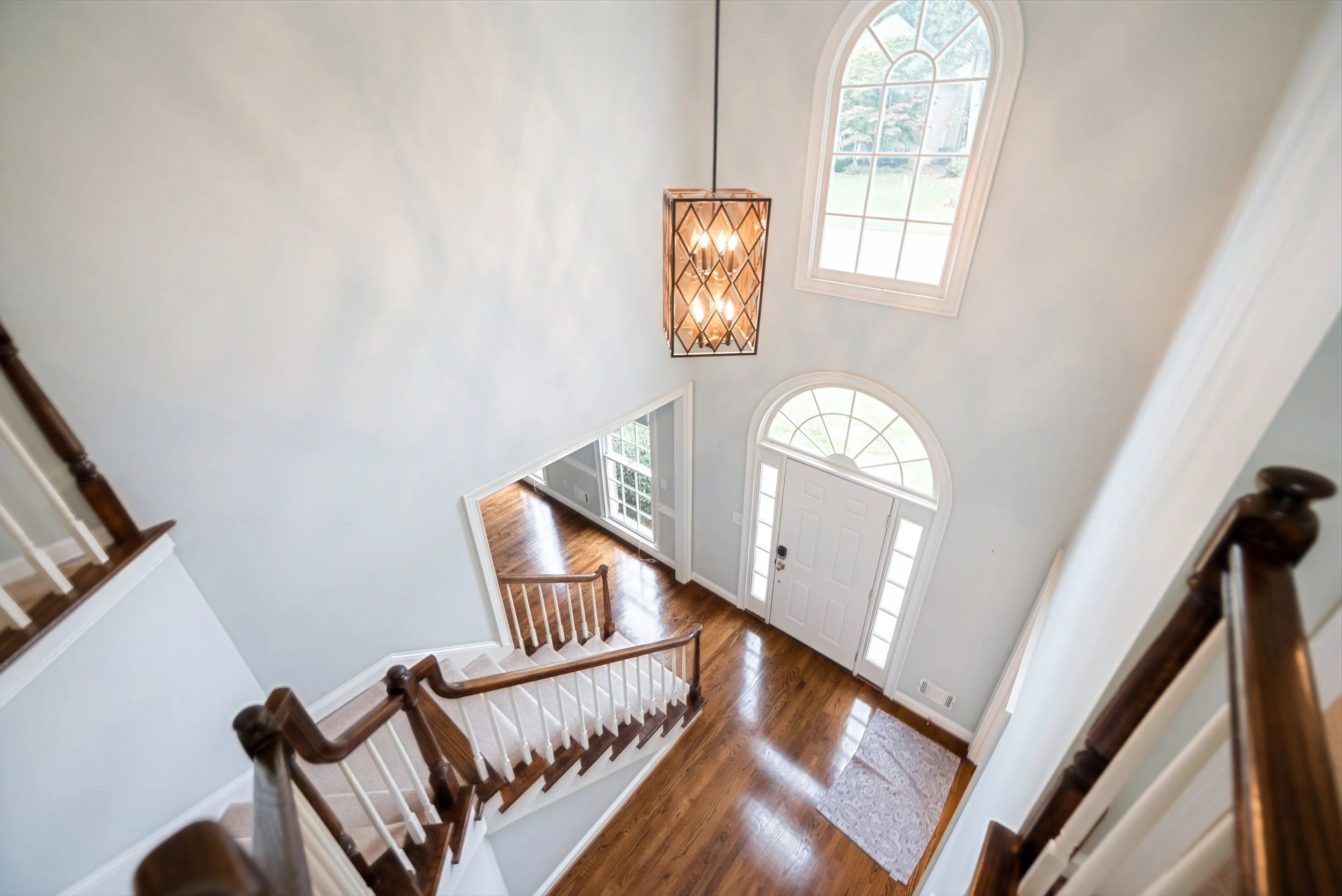 View of a bright foyer from the staircase, with hardwood floors, a large front door with windows, a hanging lantern light fixture, and two large arched windows letting in natural light.