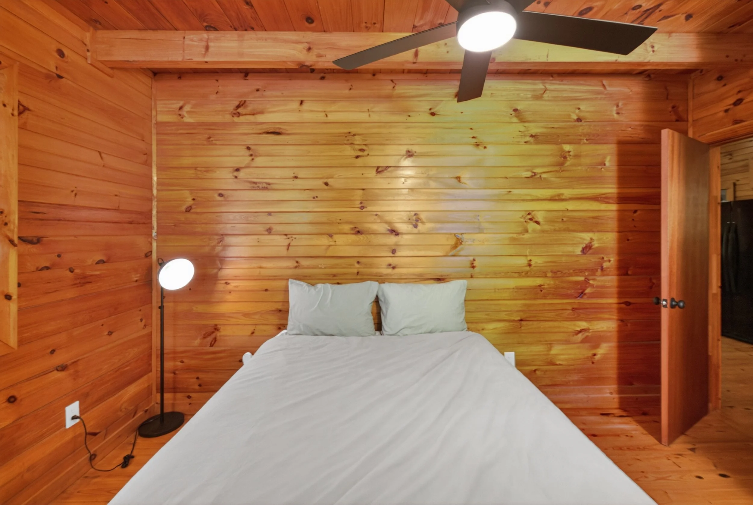 A bedroom with wooden panel walls and ceiling, a bed with white bedding and two pillows, a floor lamp with a globe light, a ceiling fan with lights, and a partially visible door.