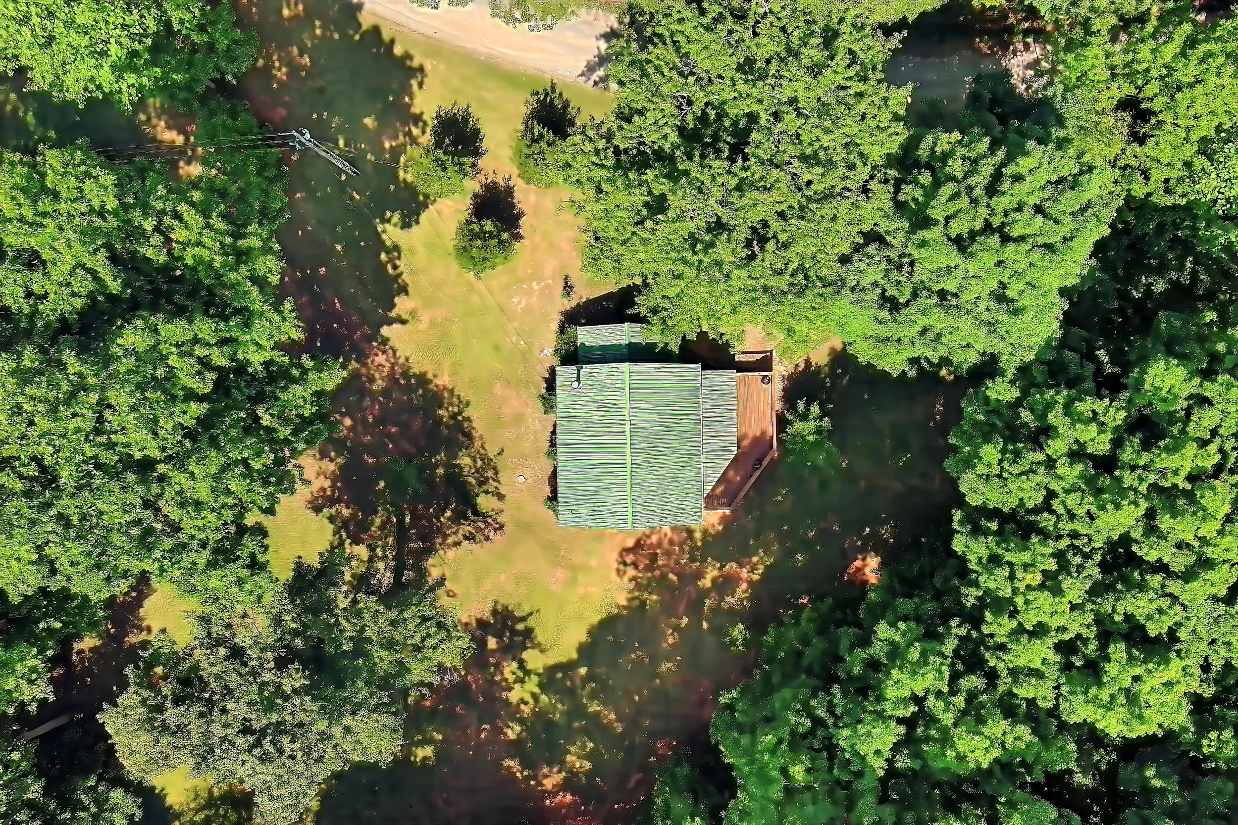 An aerial view of a house surrounded by dense green trees and grass, with a metal roof and a small deck in the backyard.