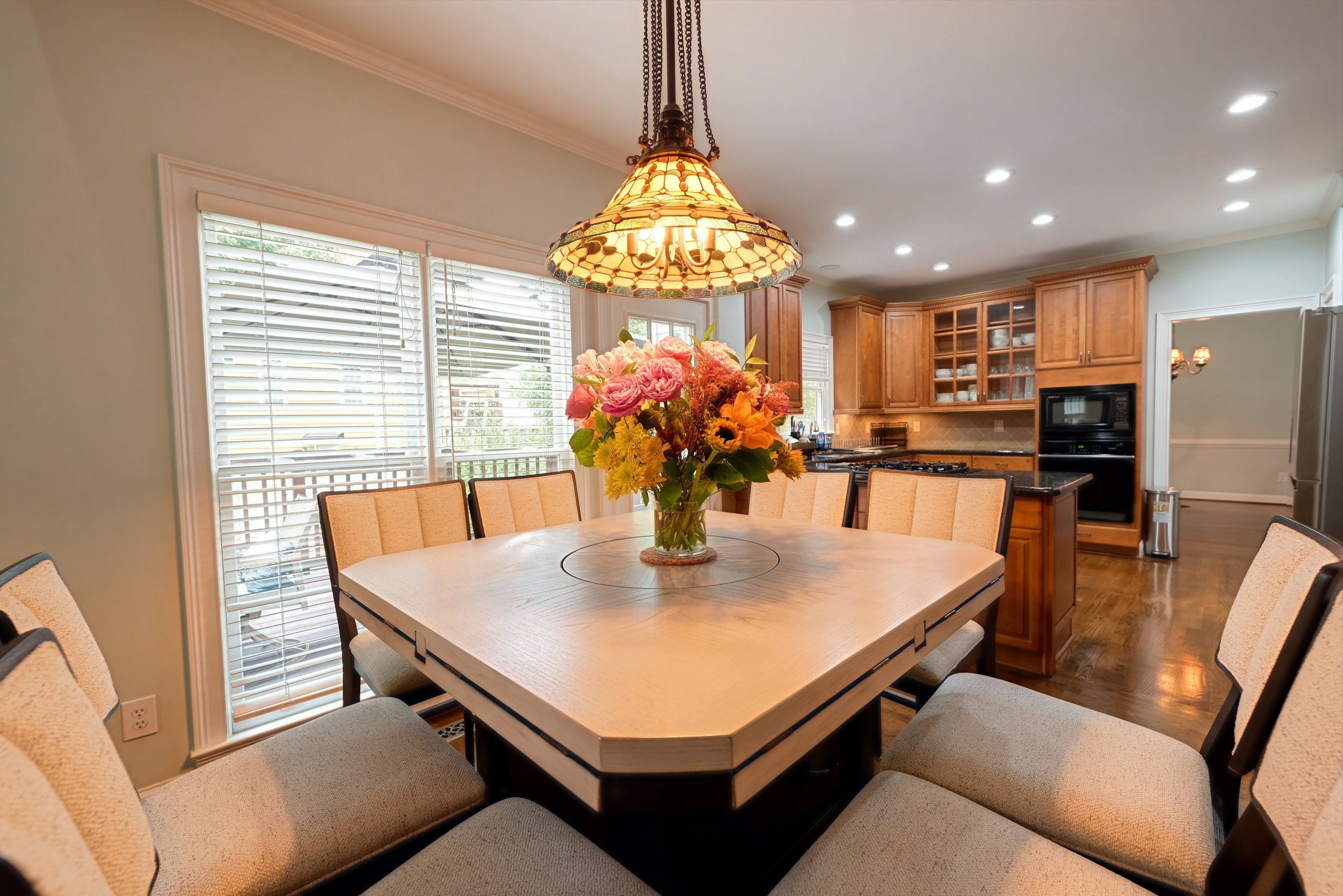 A dining room with a wooden table, beige cushioned chairs, a floral centerpiece, and a stained glass pendant lamp, with large windows, white blinds, and a view of the backyard.