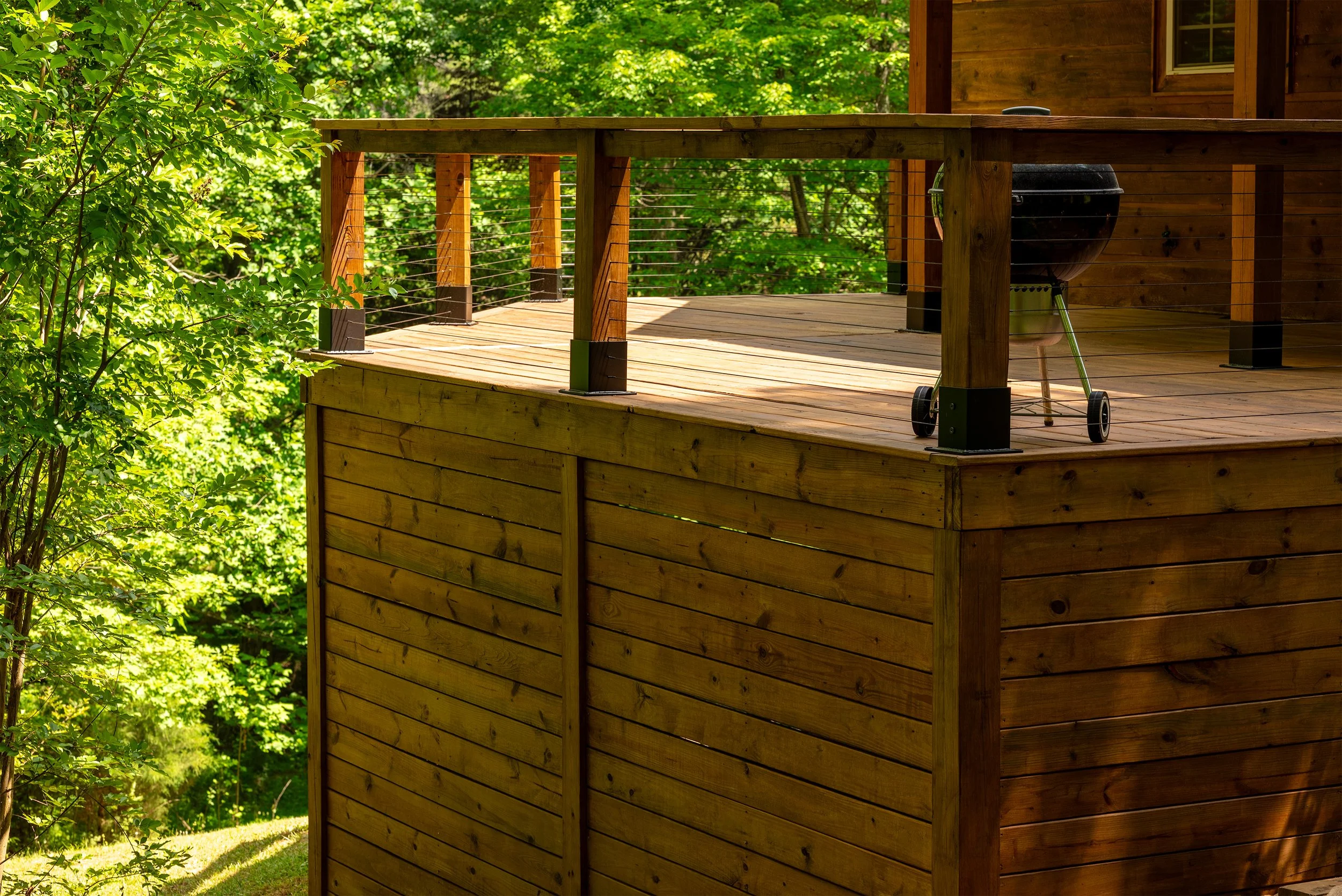 Wooden deck with railing and a charcoal grill on a house porch surrounded by green trees and foliage.