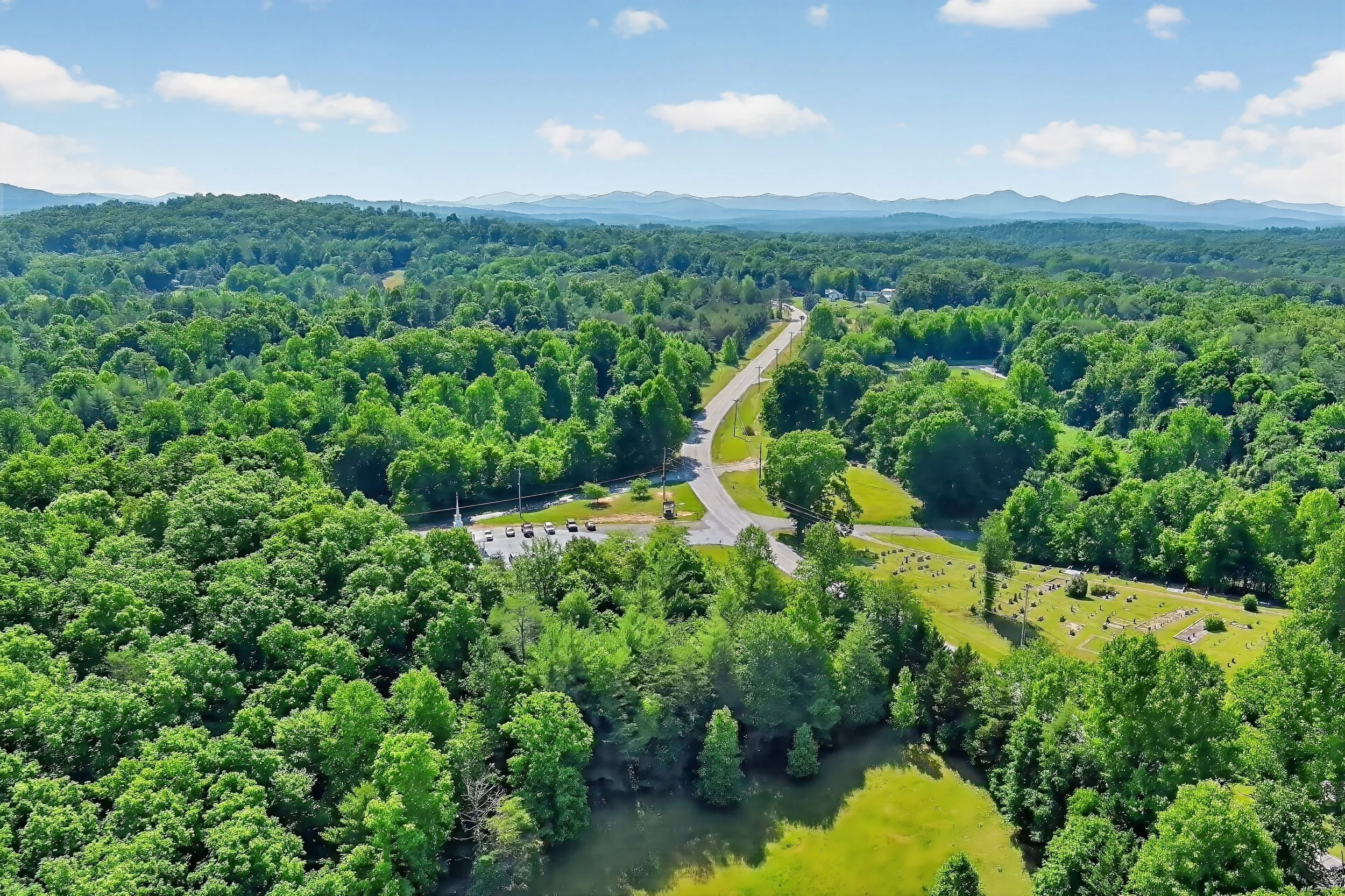 An aerial view of a lush green landscape with a winding road, a river, and distant mountains under a blue sky with some clouds.