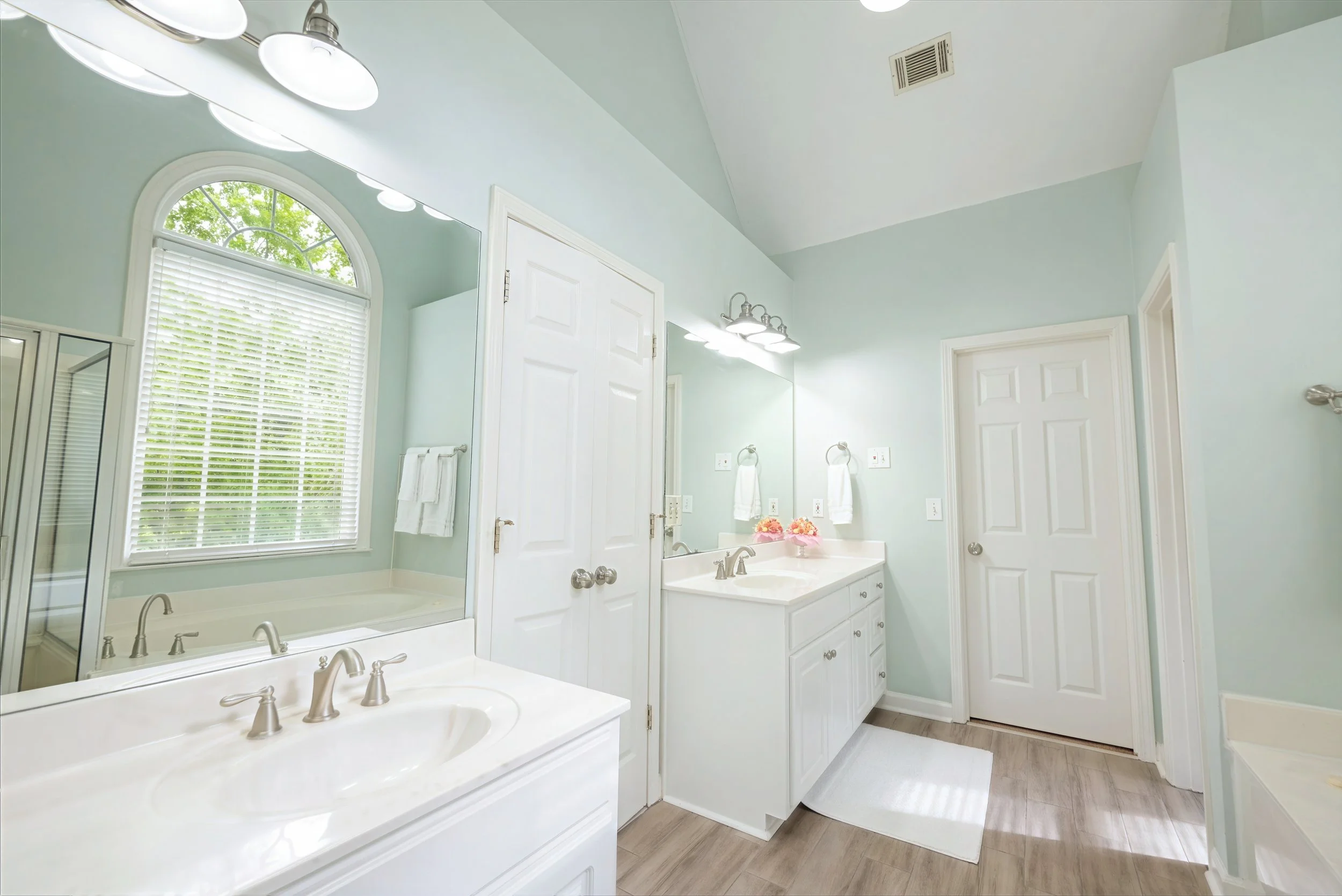 Bright bathroom with a large arched window with blinds, white cabinetry, double sinks, and modern fixtures, decorated with pink flowers on the counter.