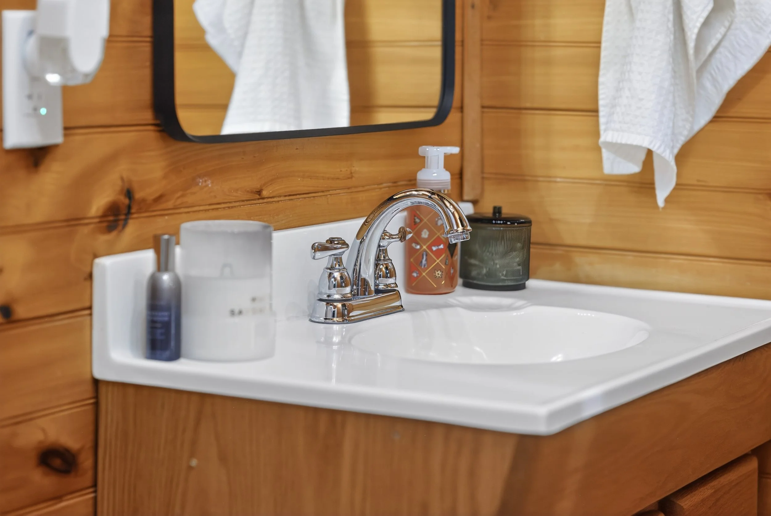 A bathroom sink with a silver faucet, surrounded by soap, a candle, and a container on a white countertop. There is a mirror, towel, heater, and wooden wall in the background.
