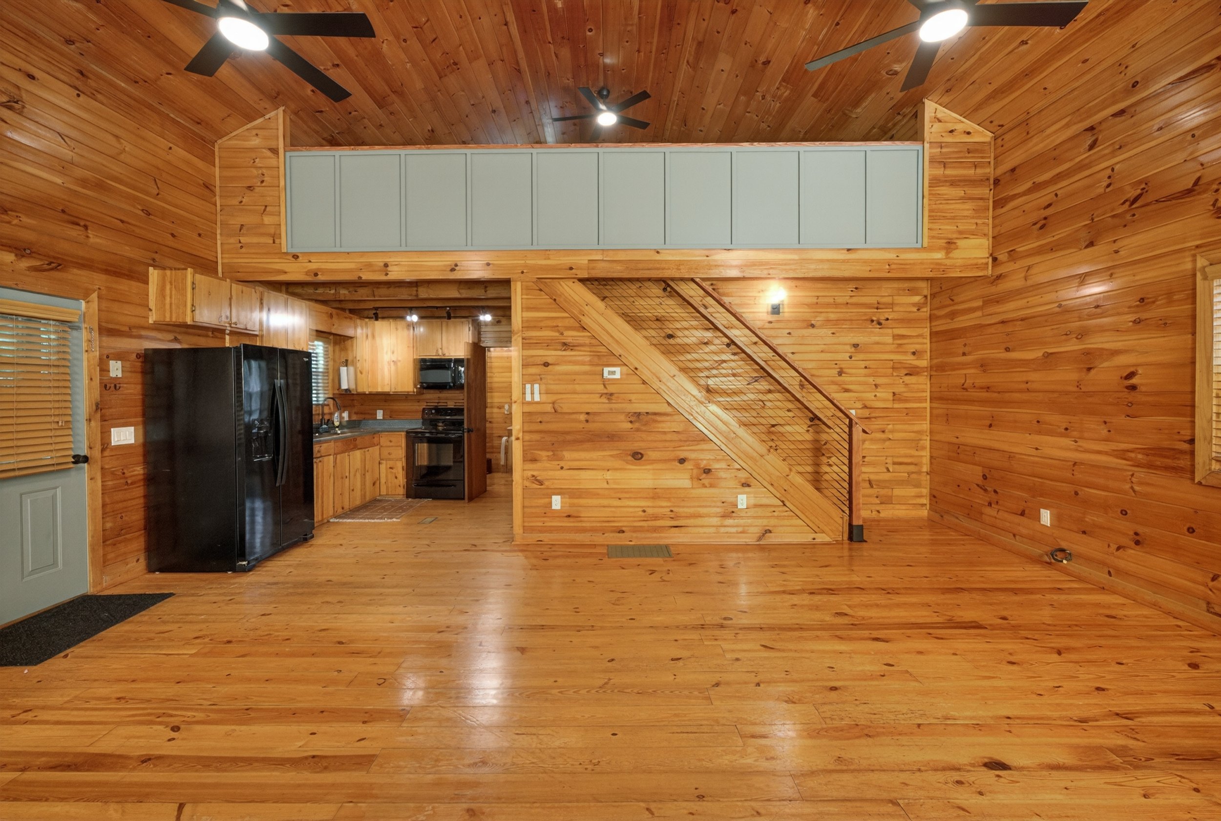 Interior of a wooden house with high vaulted ceiling, ceiling fans, and a view of the kitchen area with black appliances, wooden cabinets, and a staircase with a wire railing.