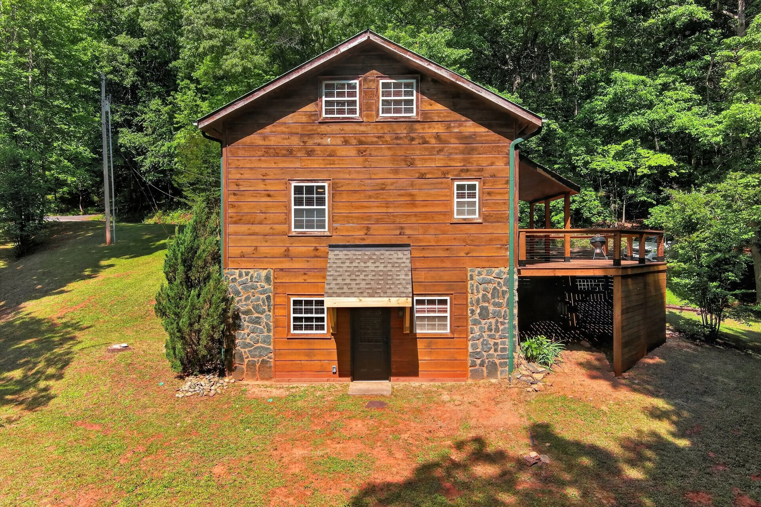 Two-story wooden house with a stone foundation, multiple windows, and a small covered porch, surrounded by green trees and grass.
