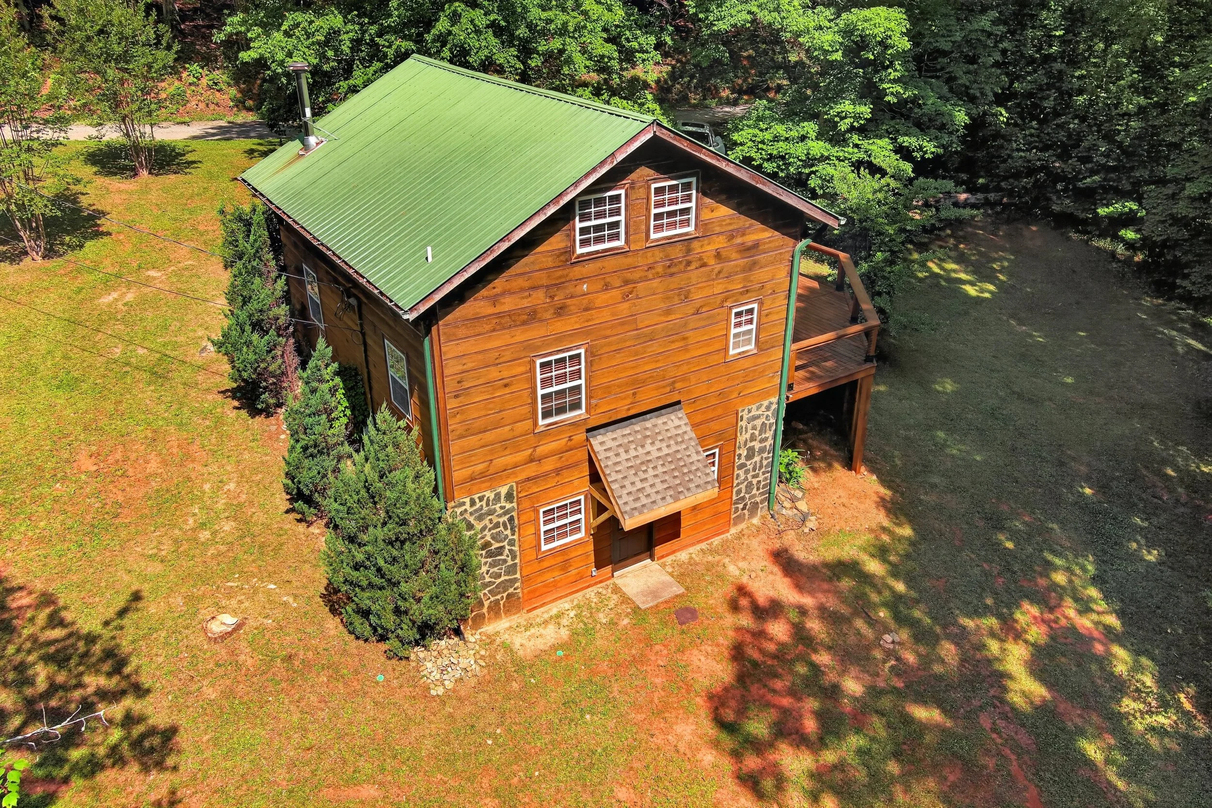 Aerial view of a two-story wooden house with a green metal roof, surrounded by trees and with a small deck, next to a pond or small body of water.