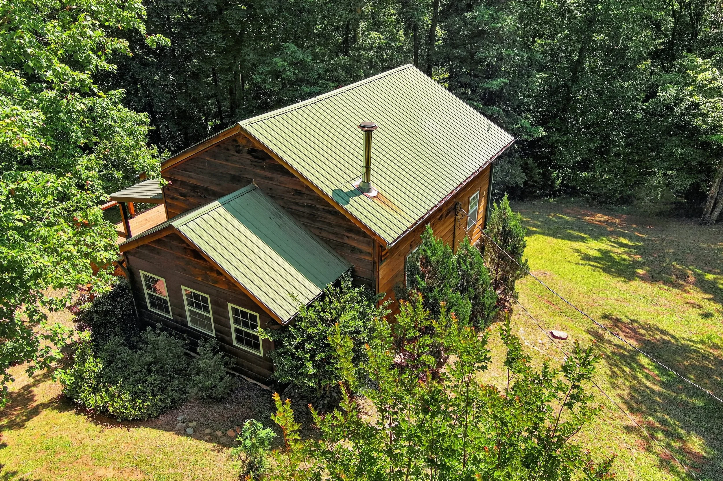 Aerial view of a wooden house with a green metal roof, surrounded by tall trees and a grassy yard in a forested area.