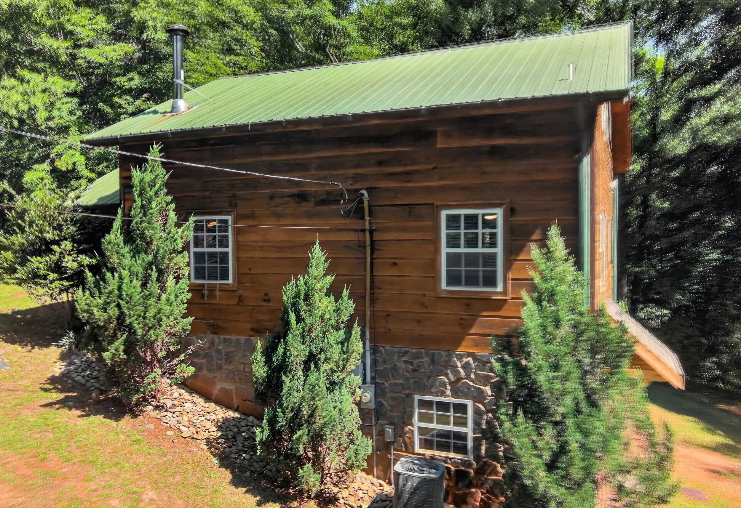 Back of a wooden house with a green metal roof, surrounded by trees and shrubs, with a stone foundation and visible roof vents.