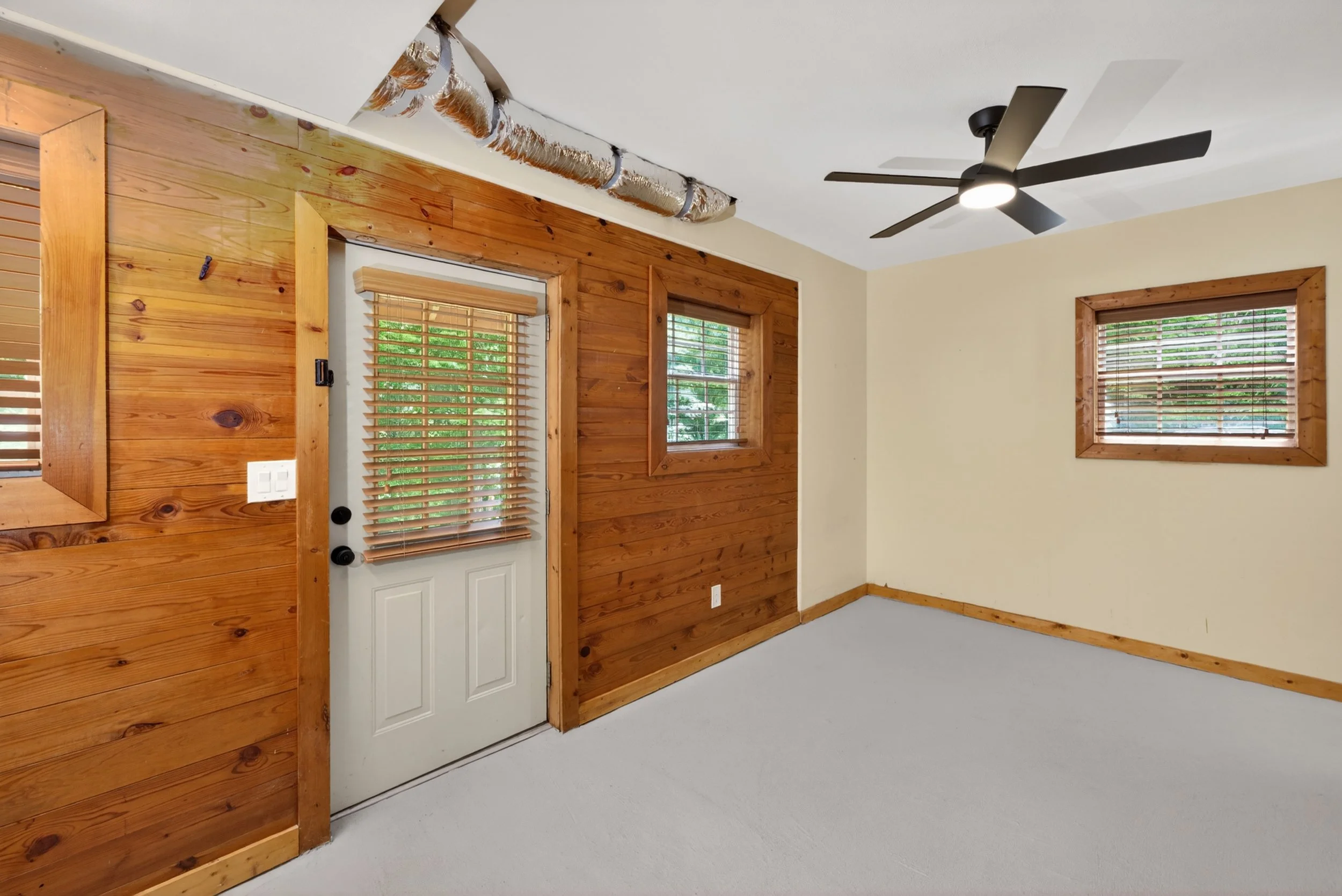 Room interior with wood paneling on walls, a white door with wooden blinds on a window beside it, double window with blinds, ceiling fan, and ductwork along the ceiling.