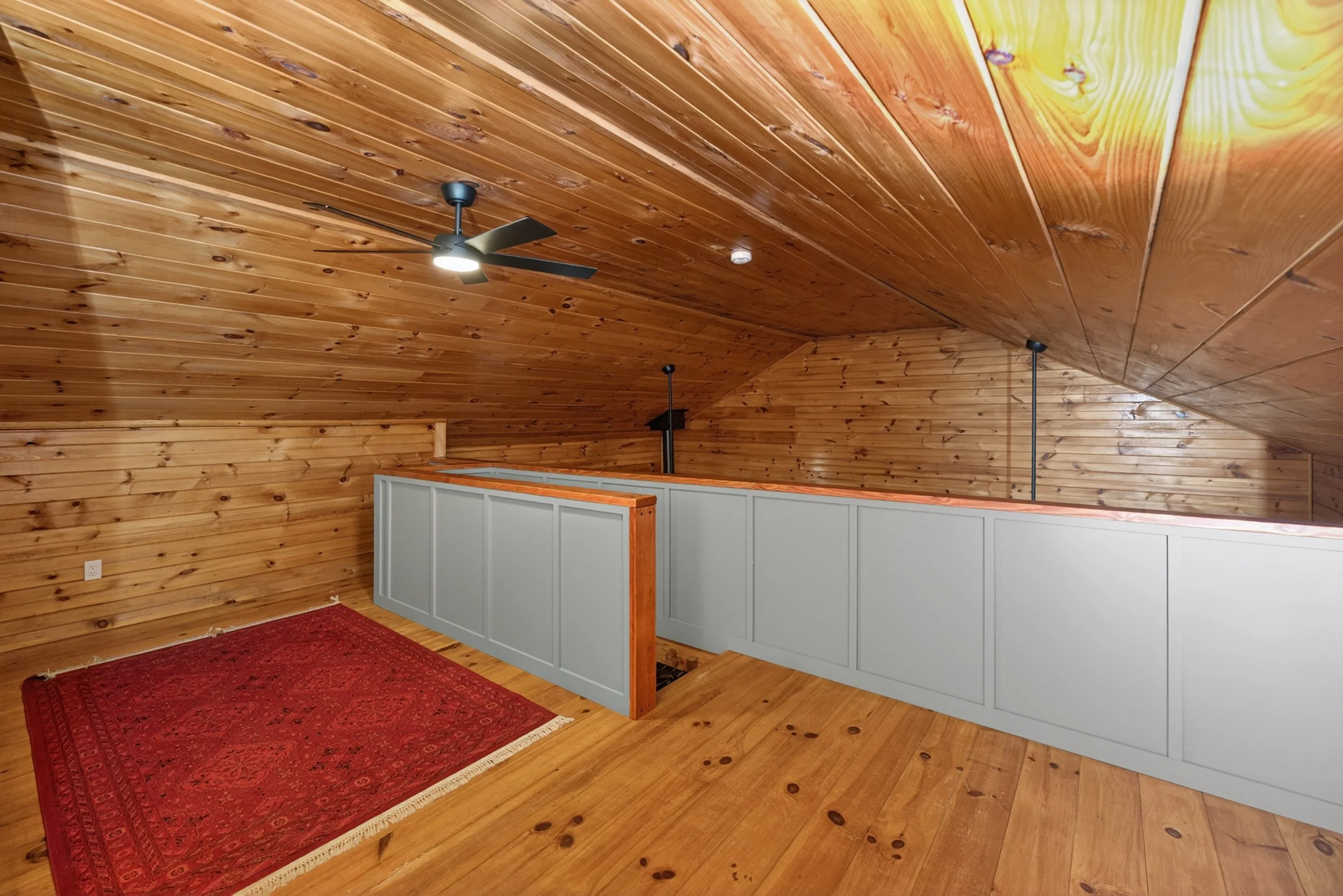 Interior view of a room with wooden paneled ceiling and walls, a ceiling fan with light, a red area rug, and a half wall with white paneling.