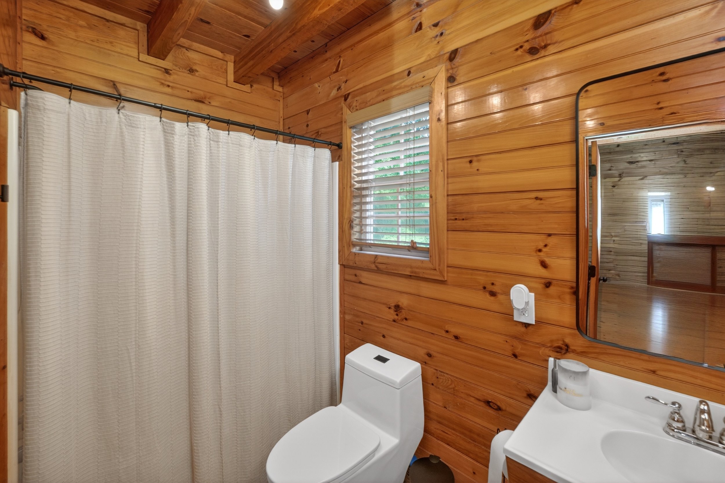 Wood-paneled bathroom with a shower with a white curtain, a window with blinds, a toilet, and a sink with a mirror.