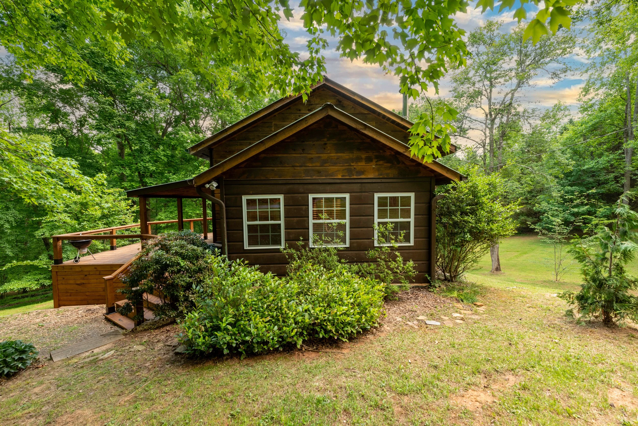 A small dark brown wooden house with white-trimmed windows is situated in a lush green forest. A wooden deck with a railing extends from the house, and a barbecue grill is visible on the deck. The scene is surrounded by trees and bushes, with a partl