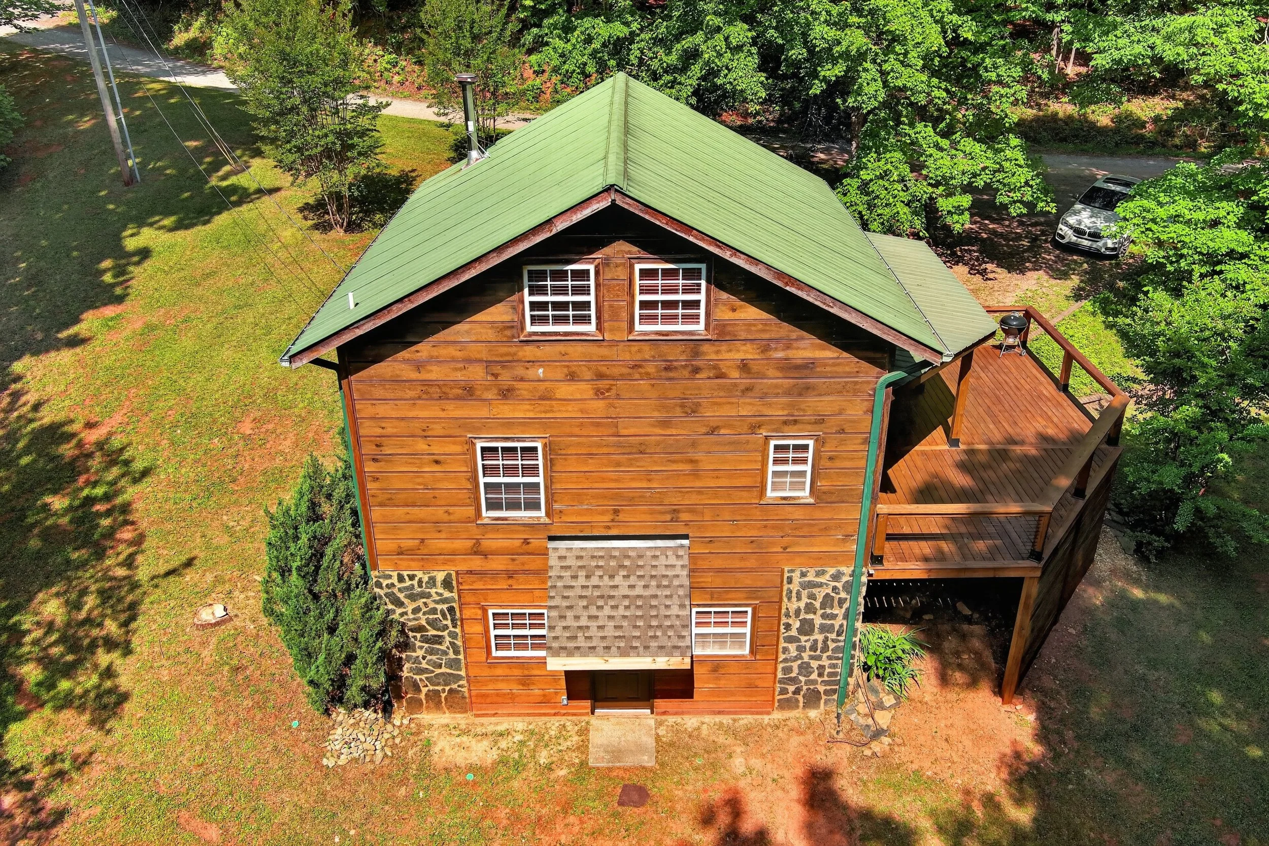 An aerial view of a two-story wooden house with a green metal roof, multiple windows, a stone foundation, and a wooden deck in the backyard, surrounded by green trees and a grassy yard, with a parked car in the driveway.