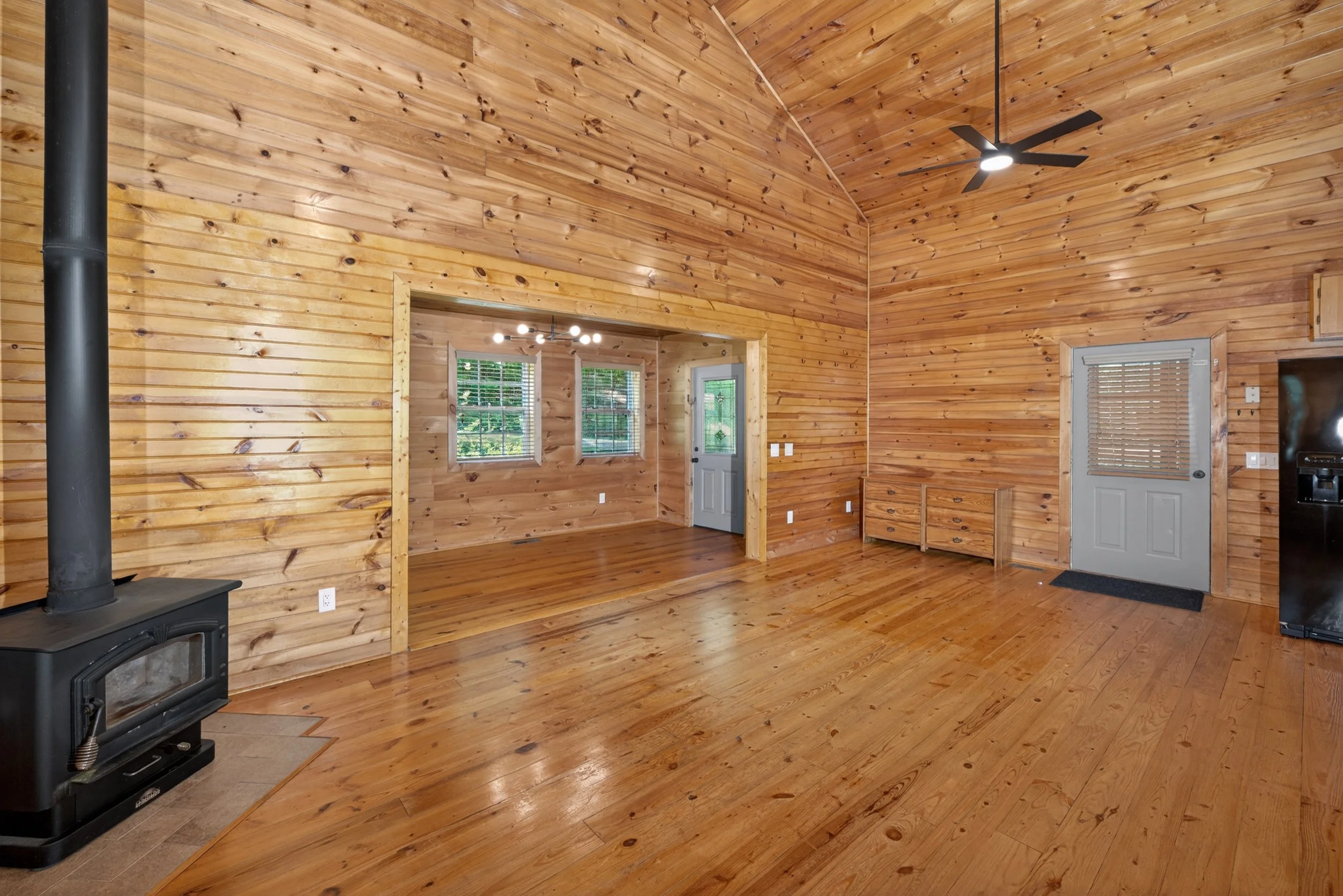 The interior of a wooden cabin with high vaulted ceilings, featuring windows, a ceiling fan, and a wood stove in the corner.