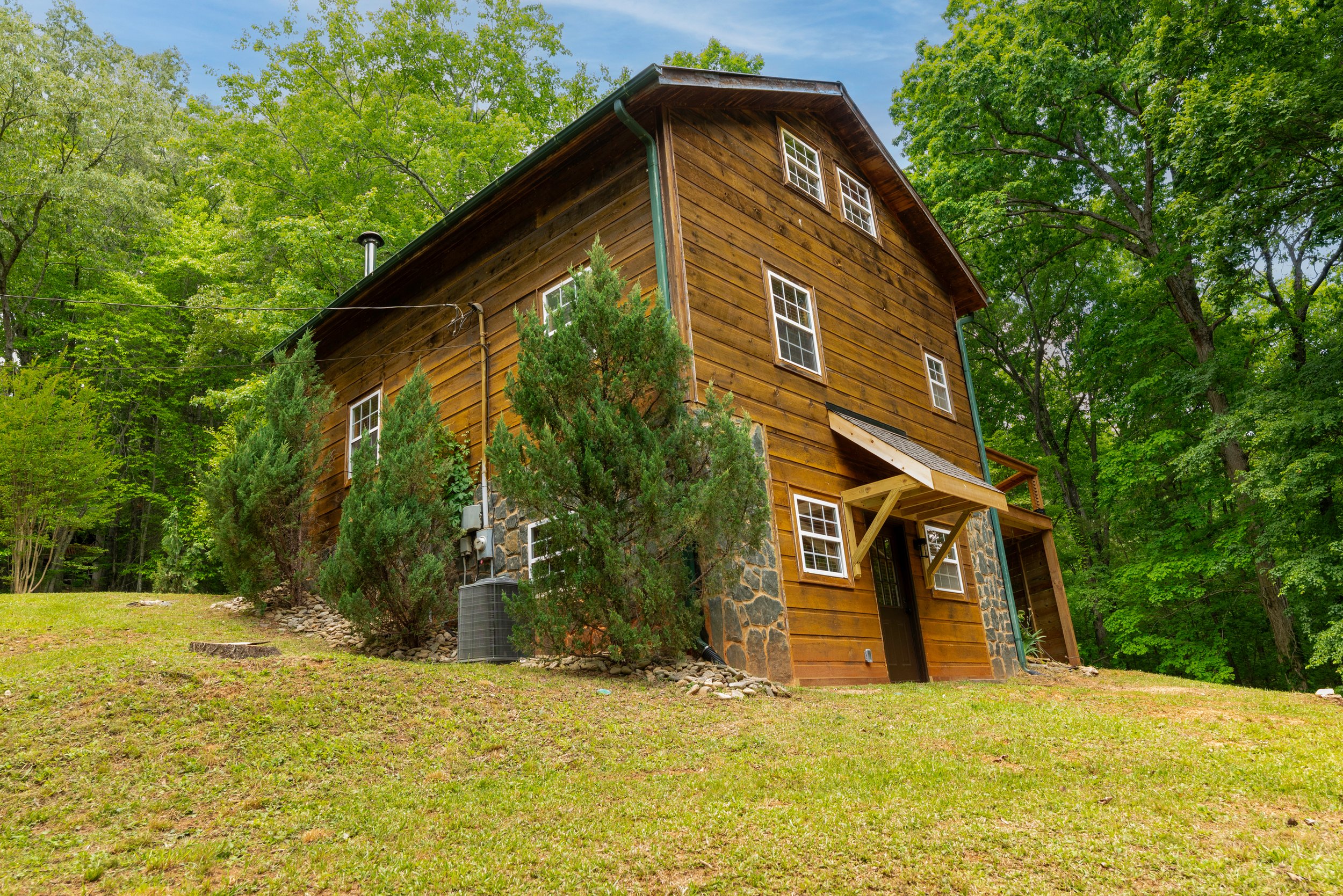 A wooden house with stone foundation surrounded by green trees and a grassy yard.
