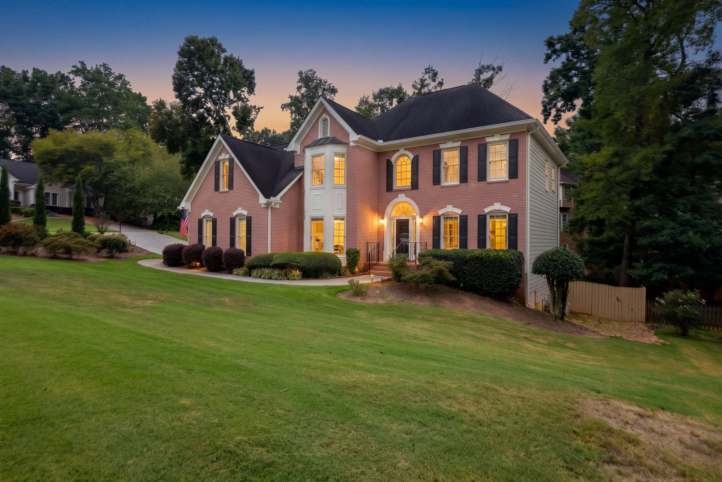 A two-story brick house illuminated from within, surrounded by a well-maintained lawn and mature trees during dusk.