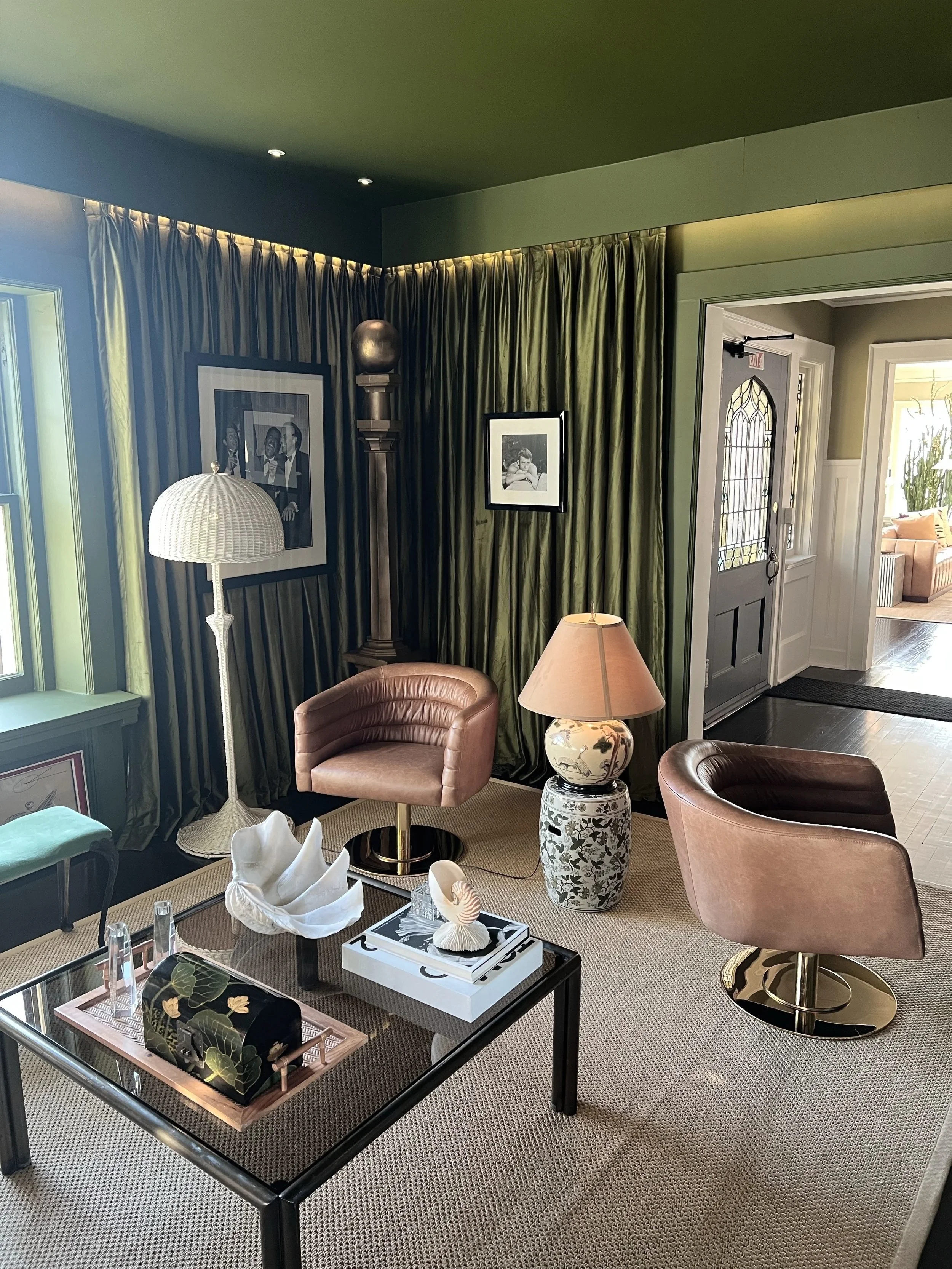 Interior of a stylish living room with pink leather chairs, a black coffee table with decorative items, a table lamp, framed photos on dark green curtains, a standing lamp, and a door leading to another room.