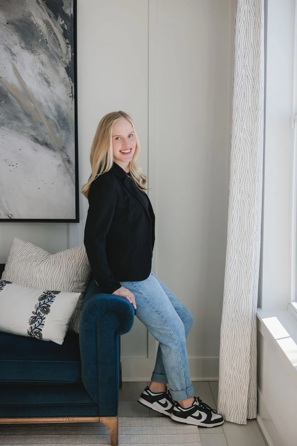 A woman with blonde hair sitting on the armrest of a blue sofa, smiling at the camera, in a brightly lit room with a large abstract painting and patterned curtains.