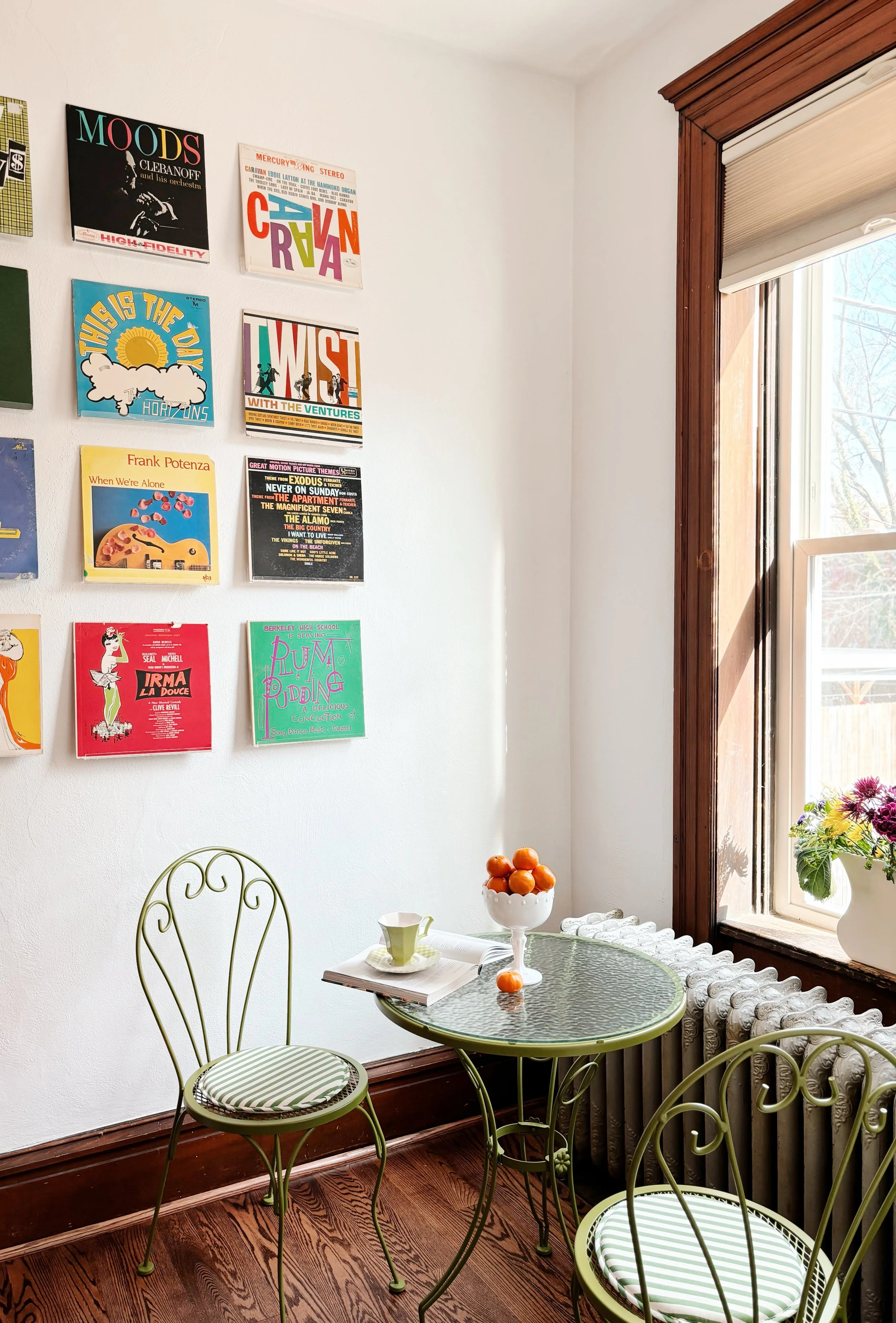 A corner of a room with a white wall decorated with colorful vintage vinyl record covers, a window with wooden trim allowing sunlight, a small green metal bistro table with two matching chairs, a white ceramic bowl filled with oranges on the table, a green mug, and a vase of flowers on the windowsill.