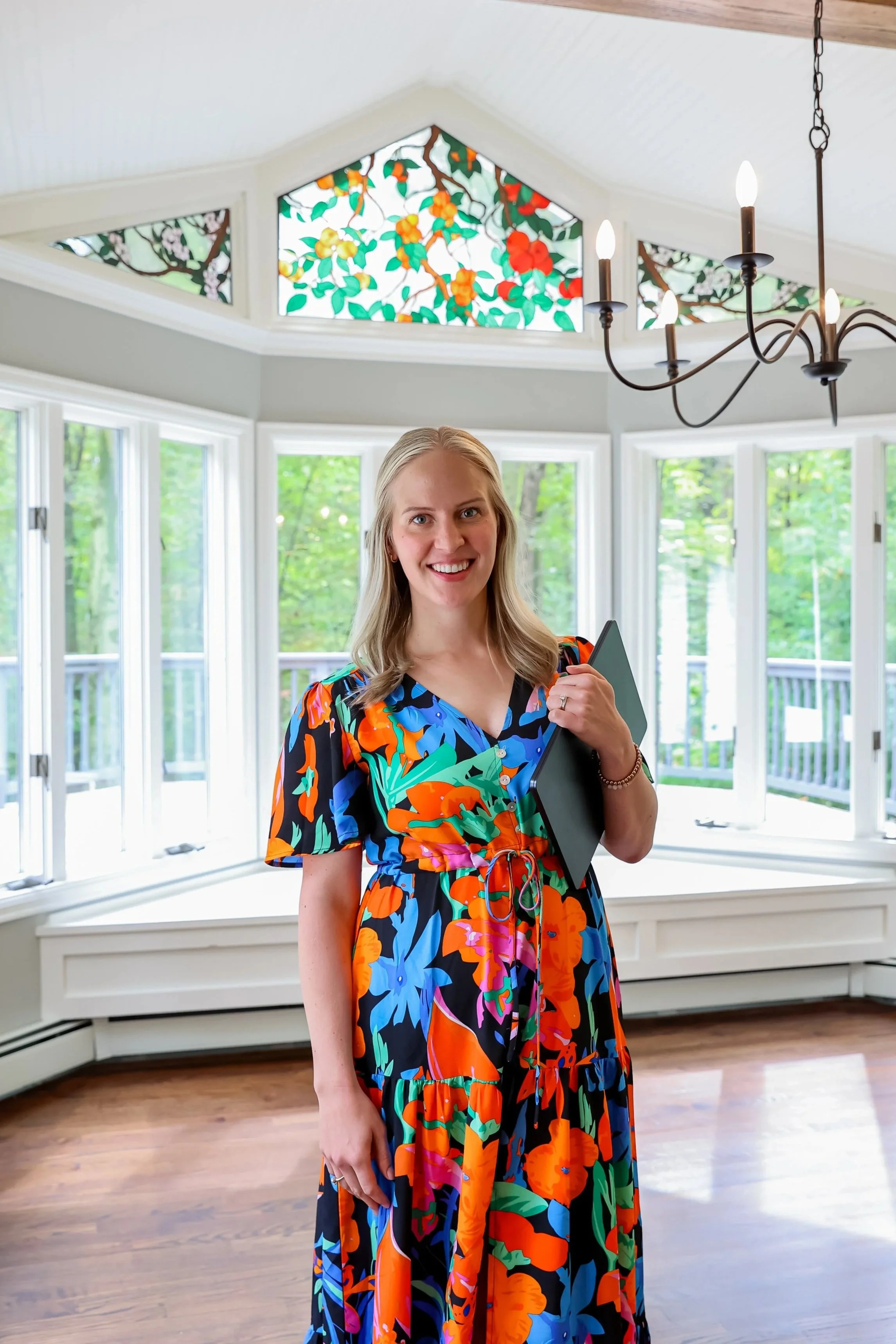 A woman wearing a colorful floral dress holding a tablet in a bright room with large windows, a stained glass ceiling, and a chandelier.