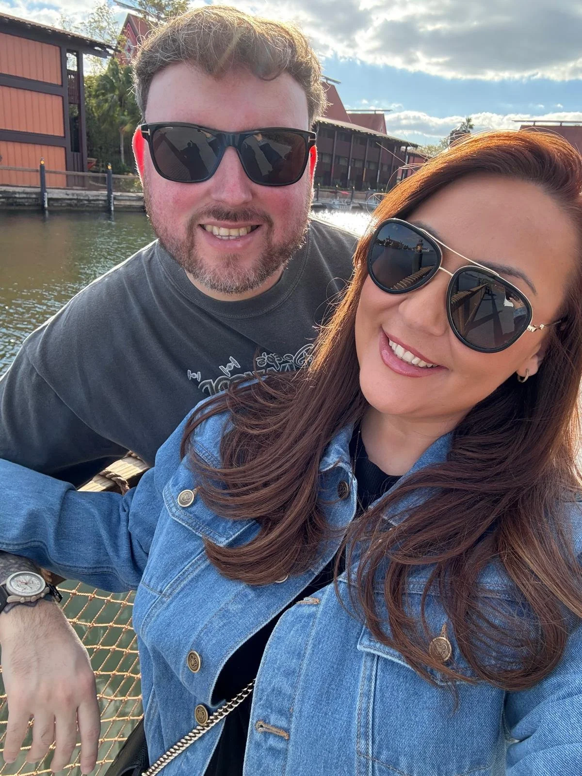 A smiling man and woman wearing sunglasses take a selfie outdoors near water, with buildings and a cloudy sky in the background.