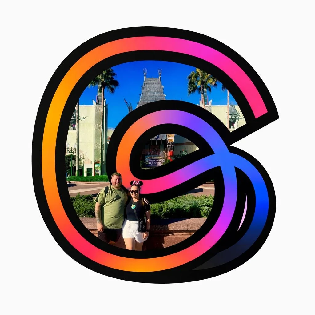 Two people standing in front of a large colorful ampersand symbol at a theme park, with a building and palm trees in the background.