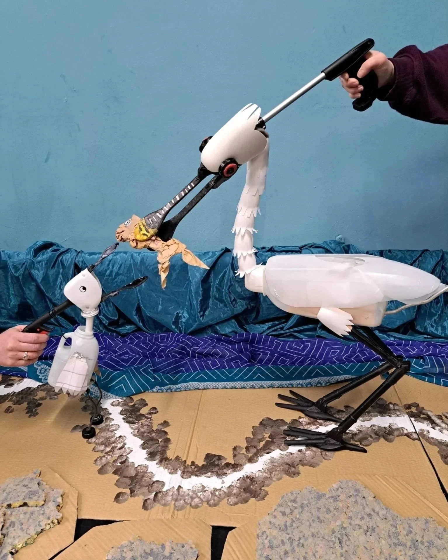 Two spoonbill puppets - an adult and its young offspring - made from household objects, against a plain blue background with a textured floor and fabric backdrop.