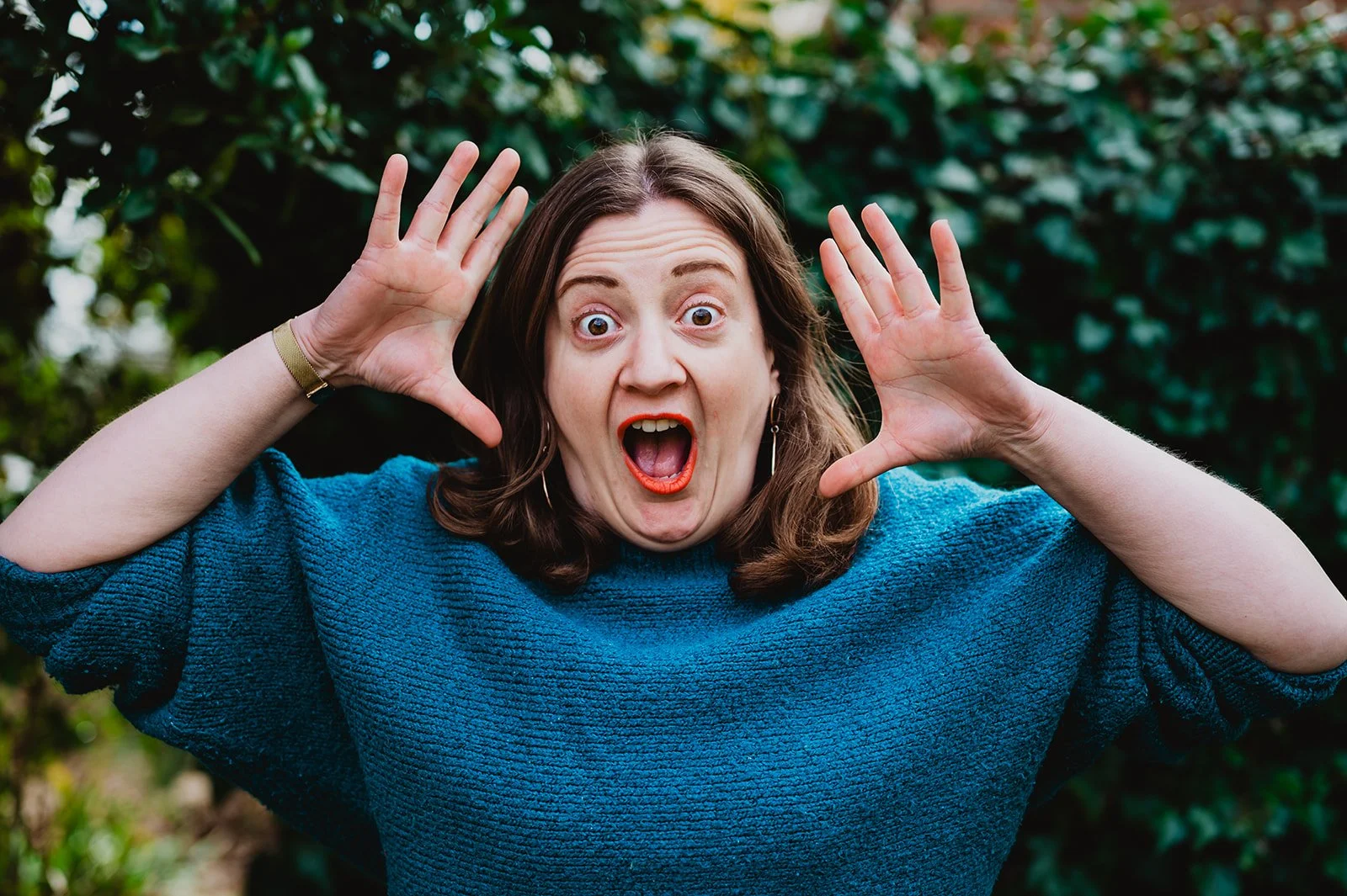Leanne, a woman with brown hair and red lipstick has her hands near her face, mouth open in surprise or shock, outdoors with green foliage in the background.