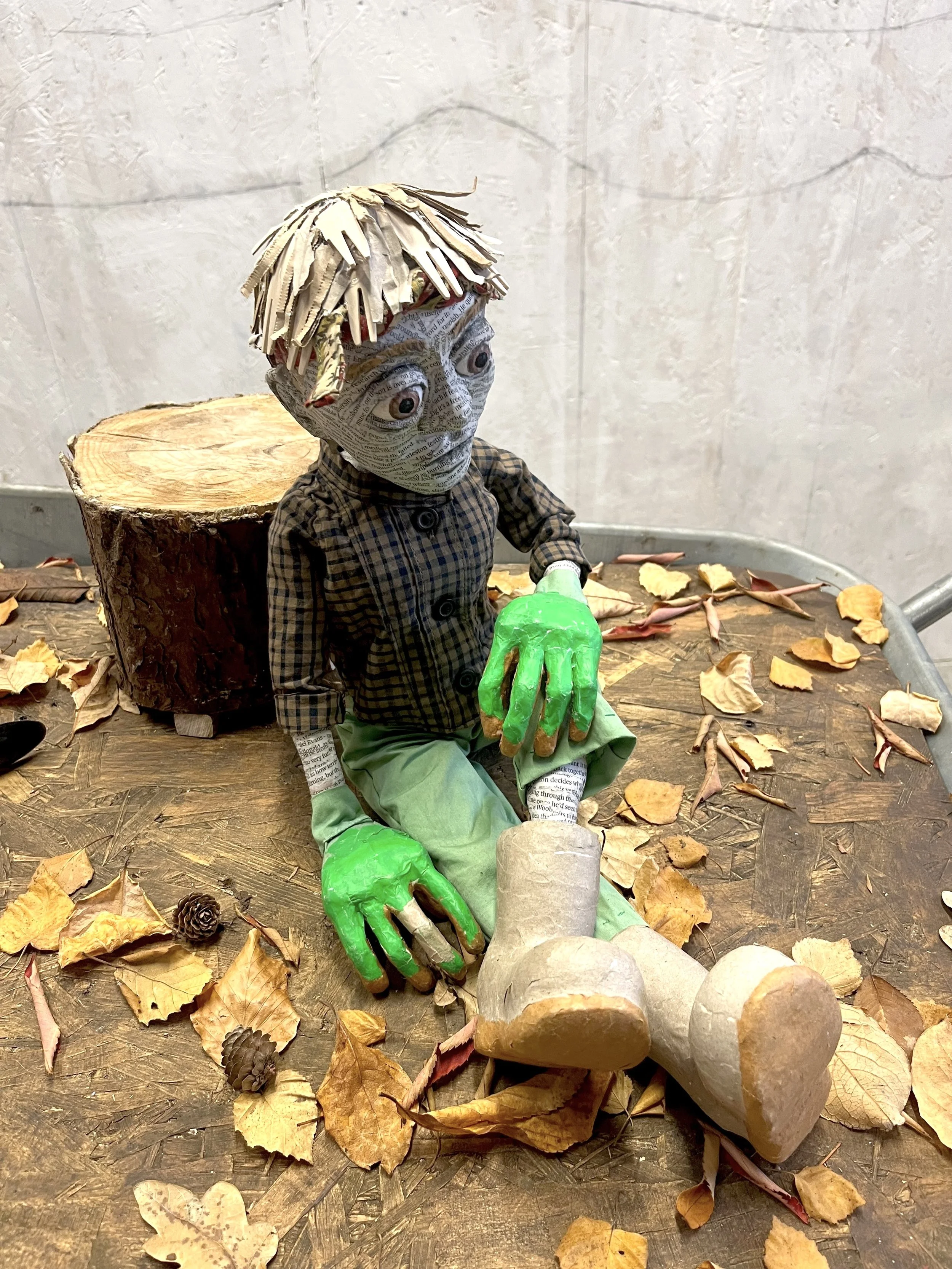A gardener puppet with green gloves, sitting on a wooden surface surrounded by fallen leaves and pinecones, with a tree stump behind them.