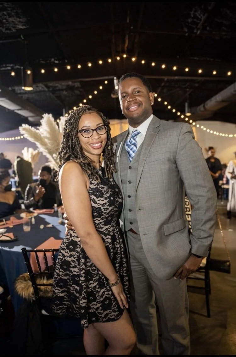 A smiling woman (India Tizol) in a black lace dress and glasses standing next to a smiling man (Antonio Tizol) in a gray suit and blue tie at an indoor event with string lights and other guests in the background.