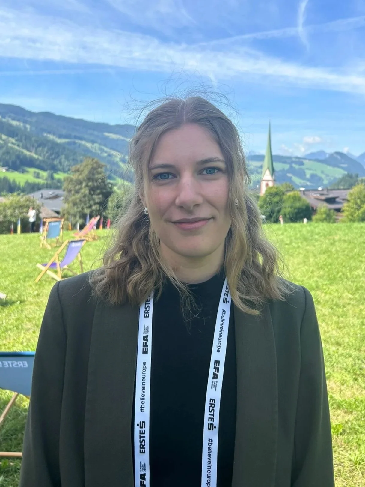 A woman with shoulder length light brown hair and blue eyes, wearing a dark jacket and a lanyard with 'ERSTI' and 'EF A' written on it, standing outdoors on a grassy field with a scenic mountain landscape, a church steeple, and colorful deck chairs in the background.