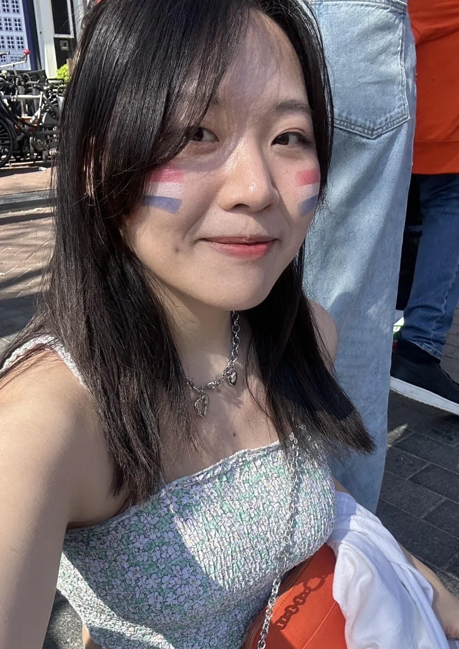 A young woman with face paint resembling the French flag, smiling outdoors on a sunny day. She has dark hair, a silver chain necklace, and is wearing a patterned dress. People and bicycles can be seen in the background.