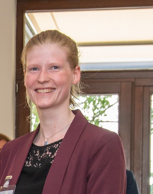 A smiling woman in a maroon blazer and black lace top, standing indoors near a window with greenery outside.