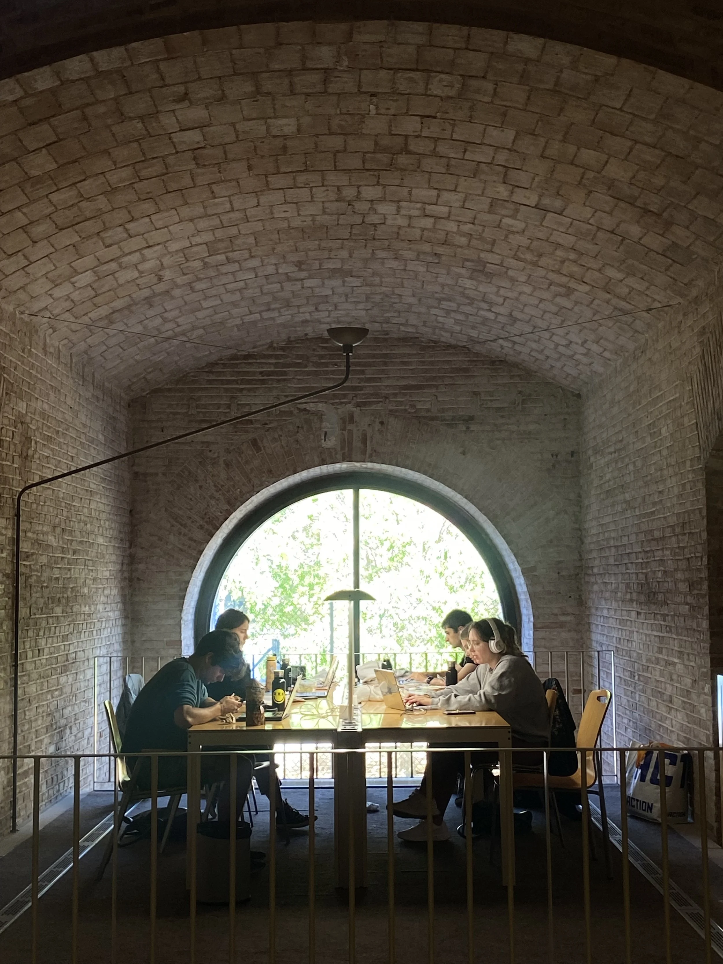 People working on laptops and using headphones at a long table inside a brick-walled room with a large round window and natural light.