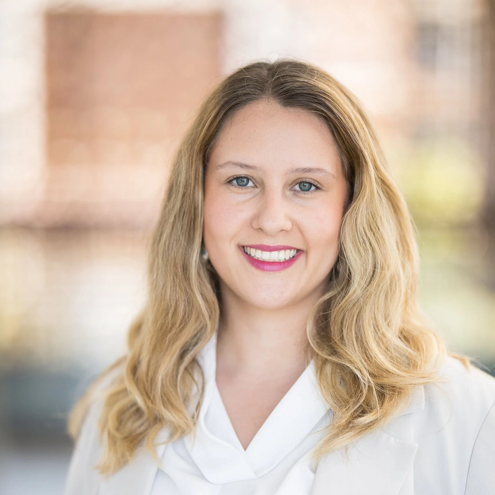 A smiling woman with long, wavy blonde hair and blue eyes, wearing a white shirt, standing indoors with blurred background.