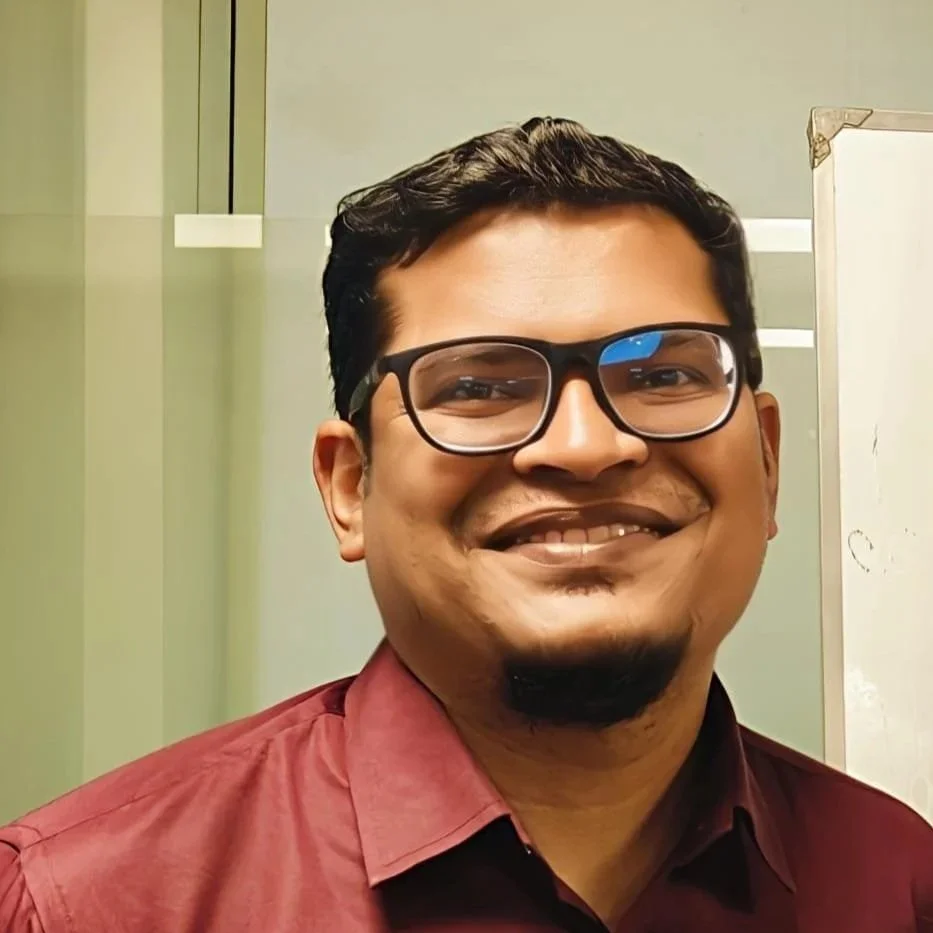 A young man with short dark hair, glasses, and light skin wearing a black shirt against a plain white background.