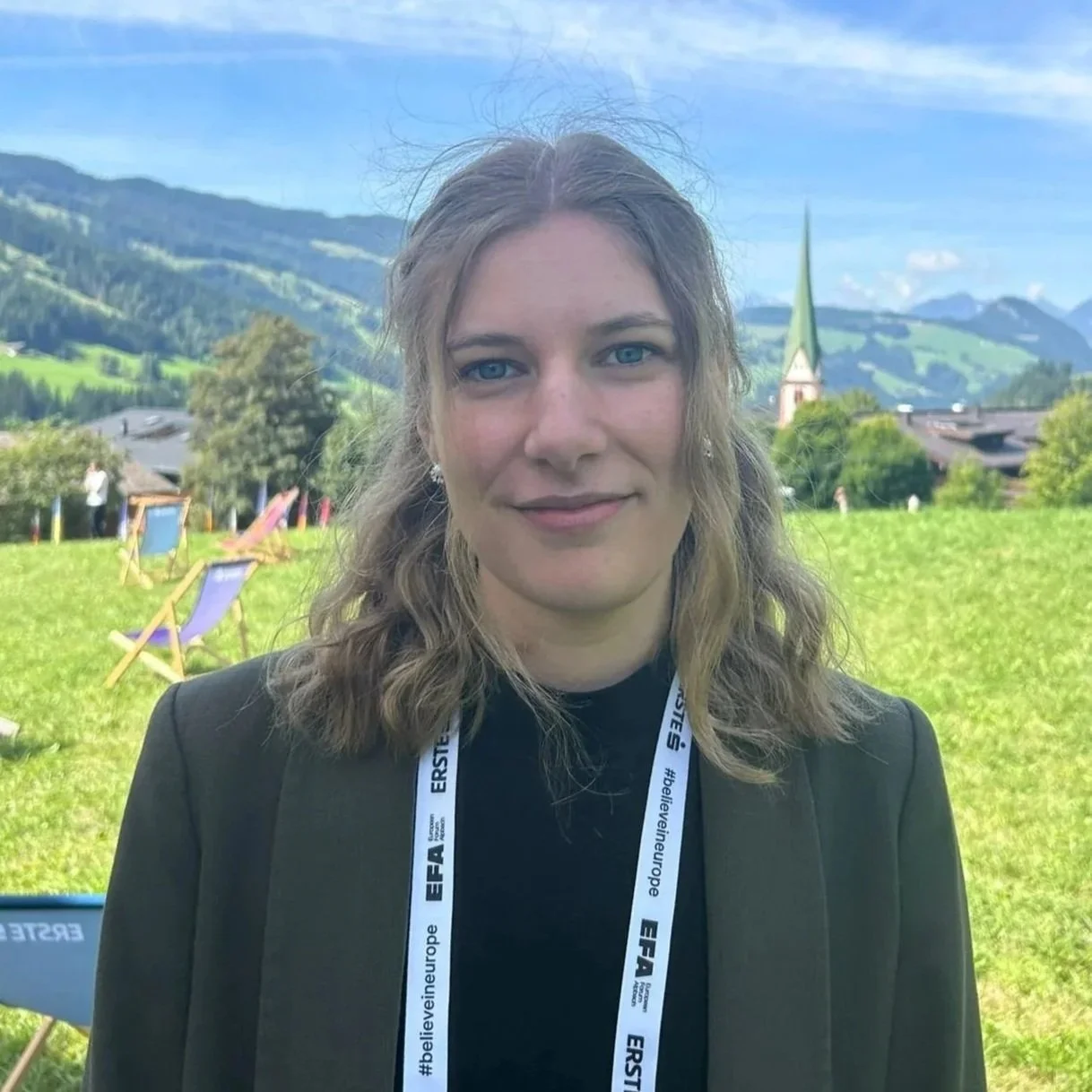 A woman with wavy, shoulder-length blonde hair and blue eyes standing outdoors in a grassy field with mountains, trees, a church steeple, and colorful chairs in the background. She is wearing a dark blazer and a white lanyard with black text.
