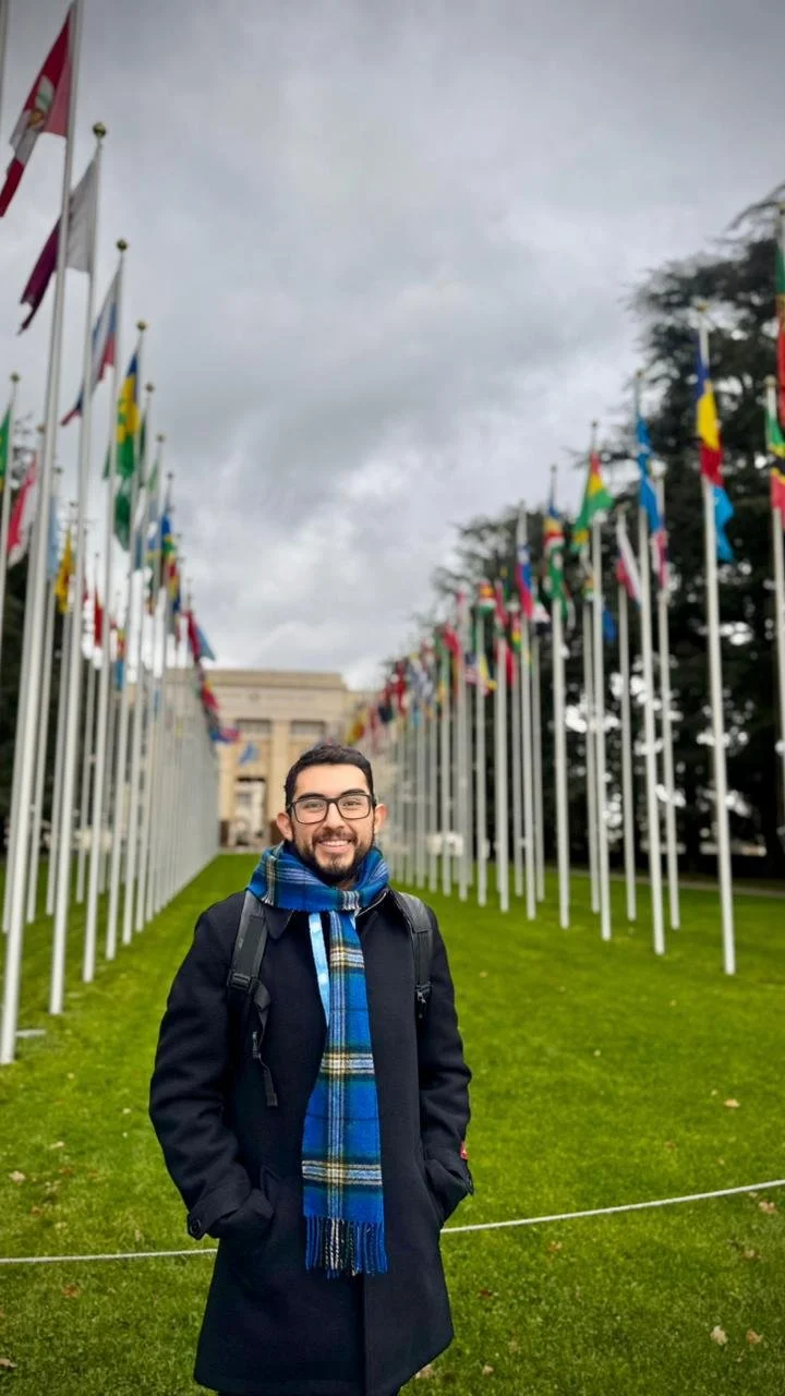 A man smiles outside a building with flags in the background.