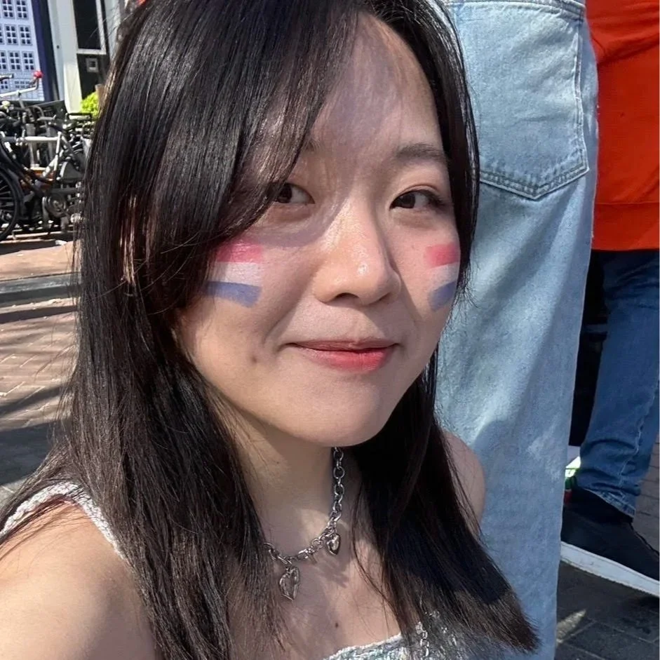 Close-up of a smiling young woman with face paint in red, white, and blue stripes on her cheeks, outdoors with bicycles in the background.