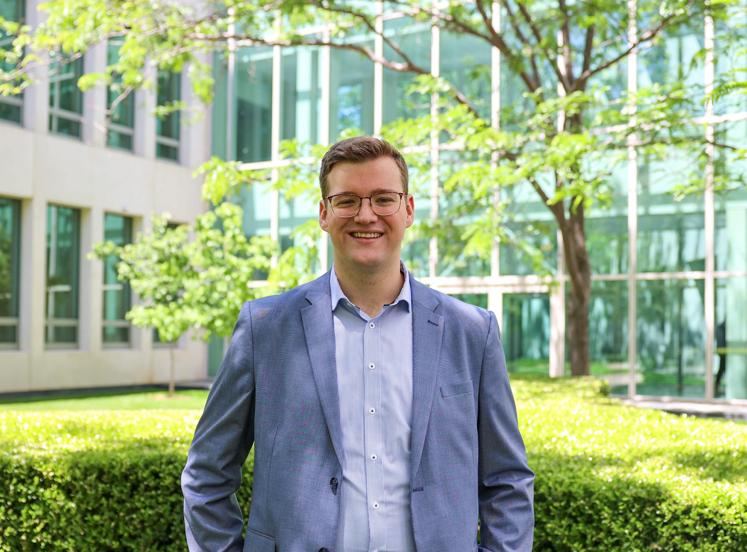 A man smiling outdoors in front of a modern glass building and green trees, dressed in a blue blazer and light shirt.
