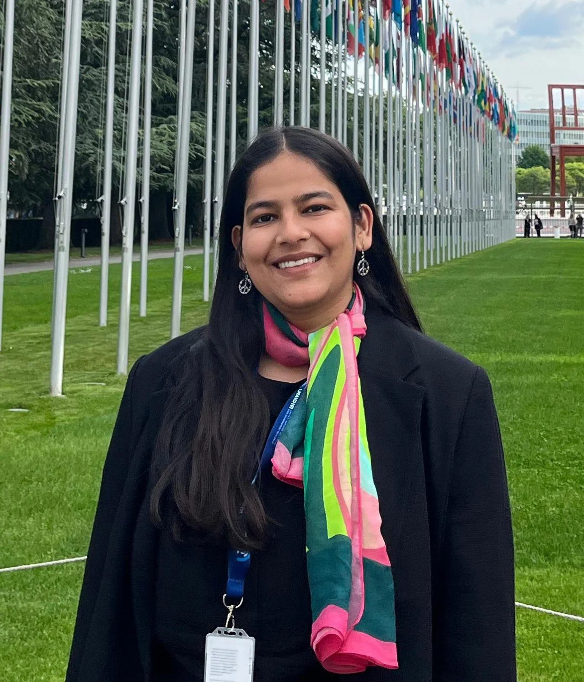 A woman with long dark hair wearing earrings, a black jacket, and a colorful scarf, smiling outdoors with grass, flagpoles, and a building with flags in the background.