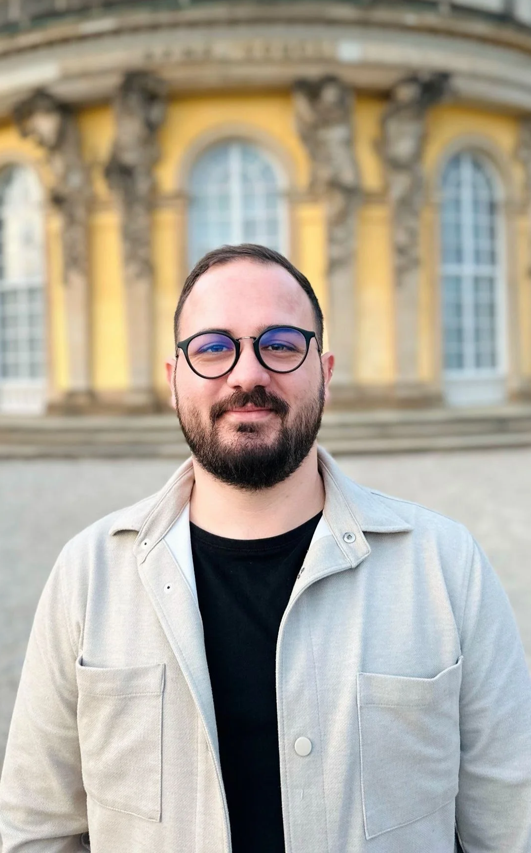 A man with glasses and a beard, wearing a light-colored jacket and black shirt, standing in front of a yellow building with large windows and ornate architectural details.