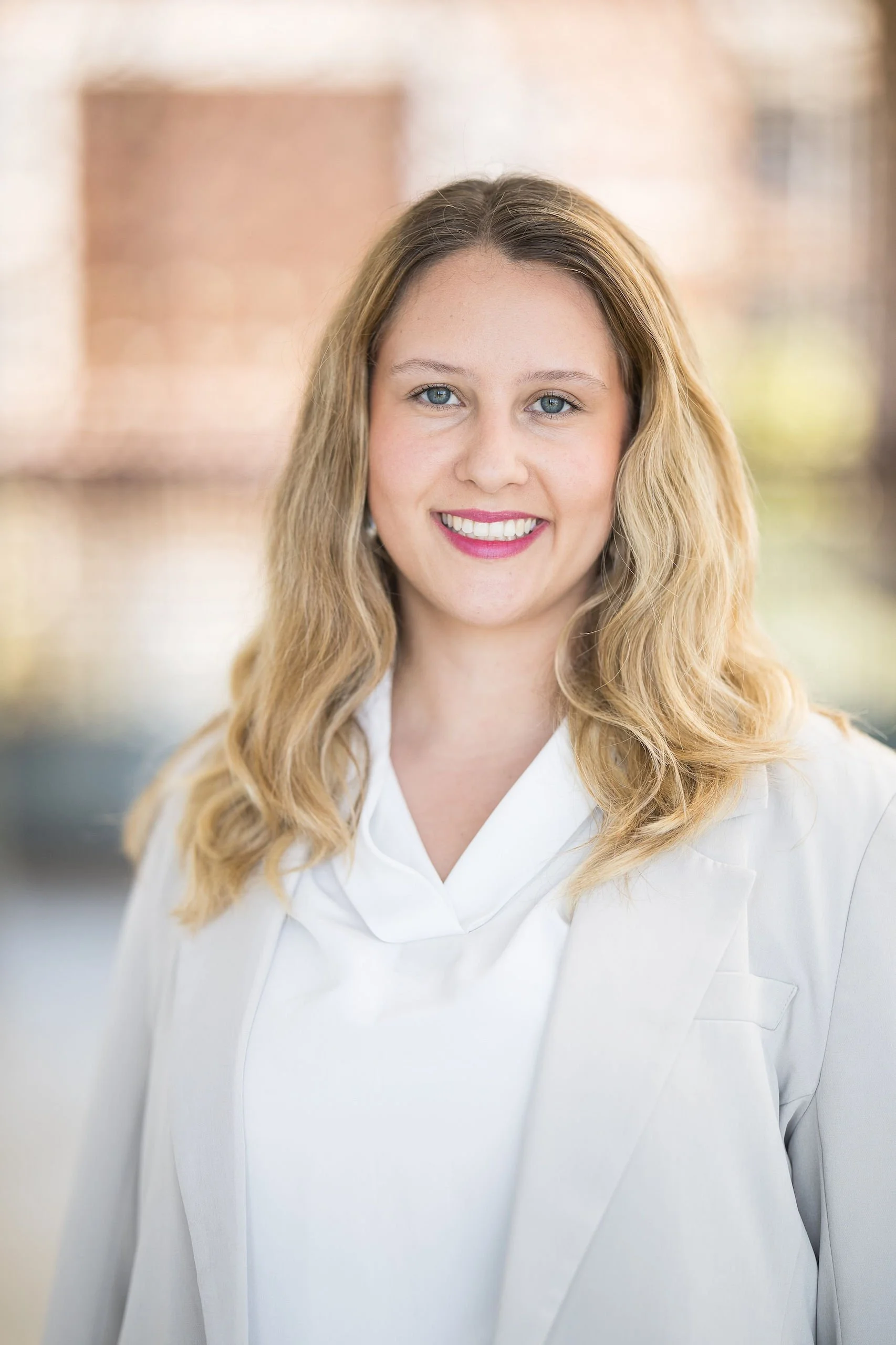 Portrait of a young woman with wavy blonde hair, blue eyes, pink lipstick, smiling, wearing a white shirt, outdoor background.