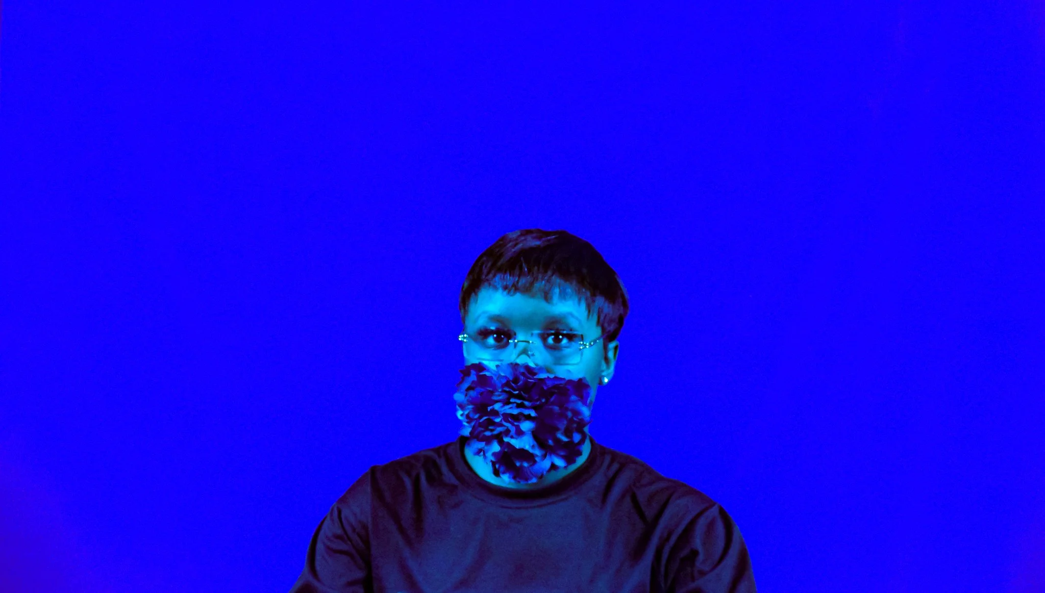 A woman with short dark hair, glasses, and earrings wearing a face mask with blue and purple flowers against a blue background.